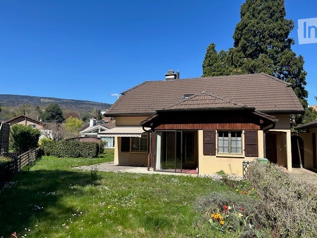 Single-story house with a brown tiled roof, yellow exterior walls, and large windows. The house has a covered porch or balcony area and a detached garage on the property. The house is surrounded by a well-maintained lawn and garden with various plants and