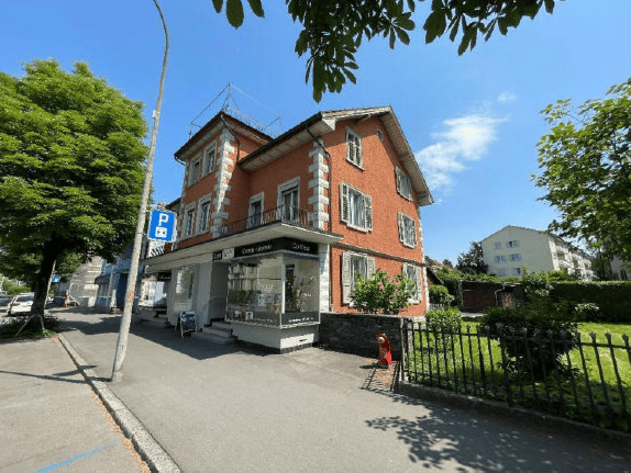 Two-story brick building with a storefront on the ground floor, surrounded by trees and a small fenced-in area.