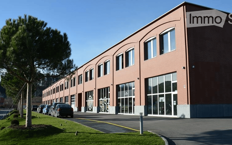 A multi-story commercial building with a brick facade, large windows, and a parking lot in front. The building appears to be a modern office or retail space surrounded by trees and landscaping.