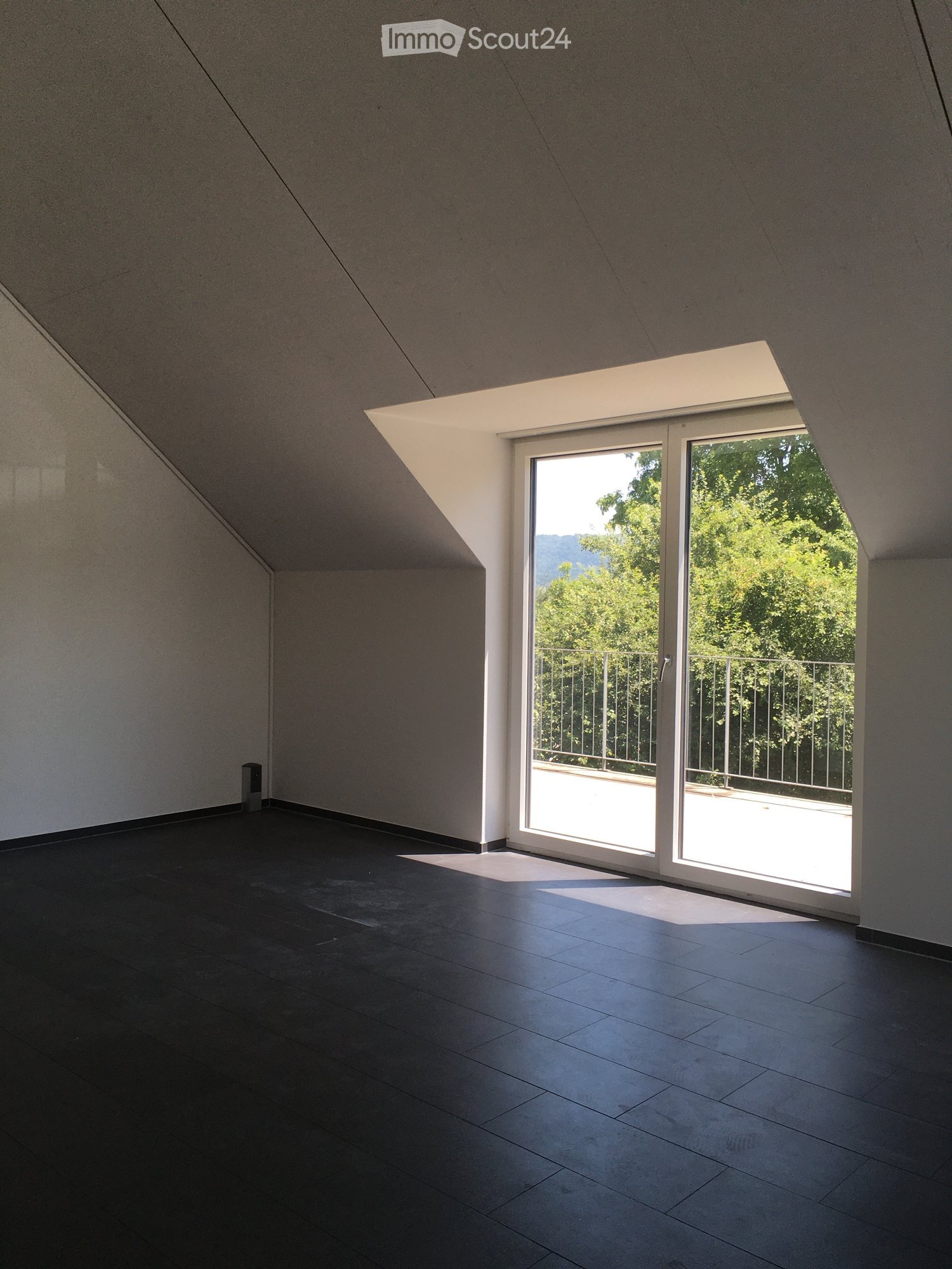 Spacious room with large windows overlooking a balcony and lush greenery outside. The room has dark tile flooring and plain white walls.