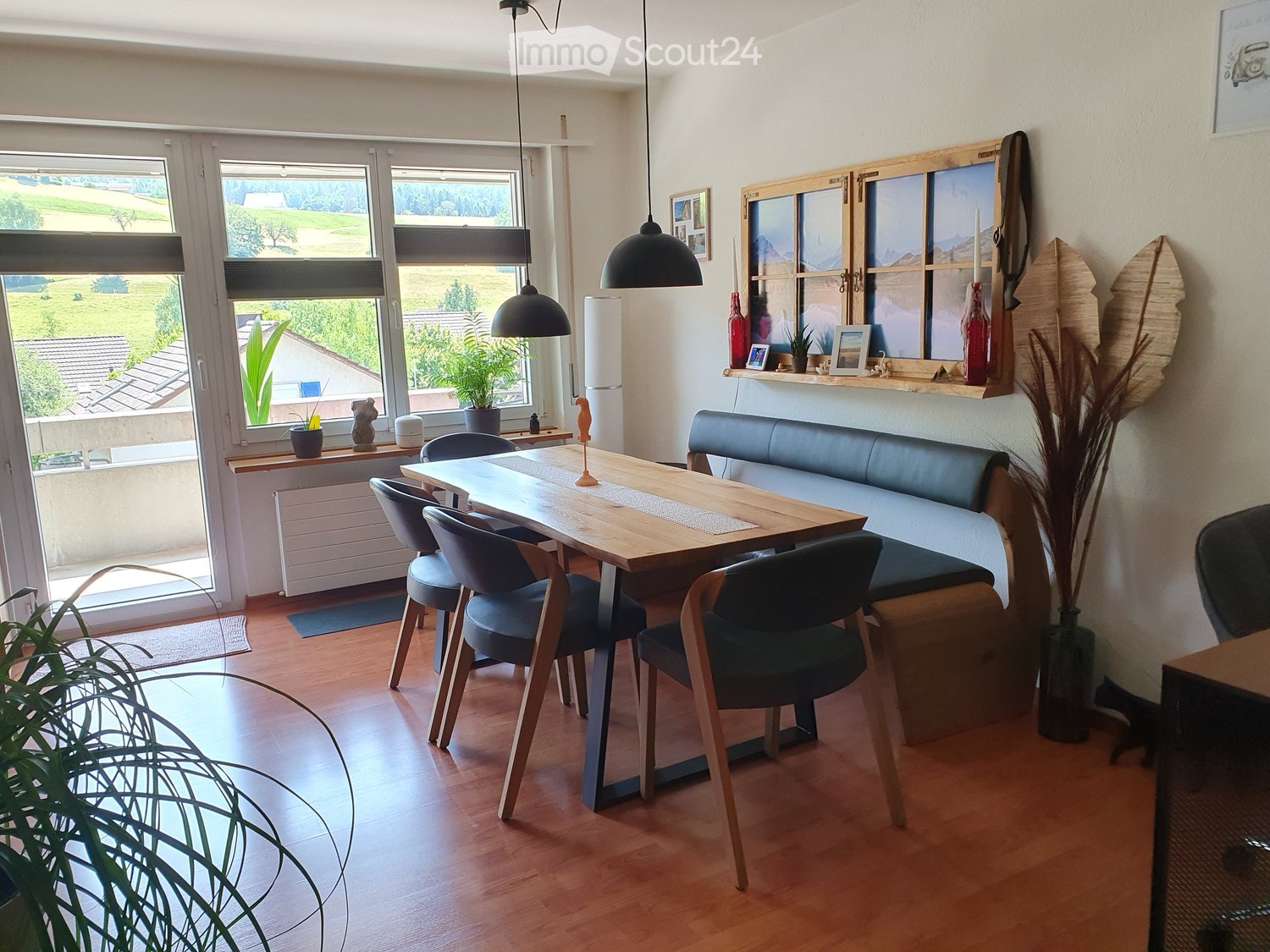 Spacious dining area with large windows overlooking a scenic outdoor view, wooden dining table and chairs, pendant lighting fixtures, shelving unit with decor, and a cozy seating area with a sofa.