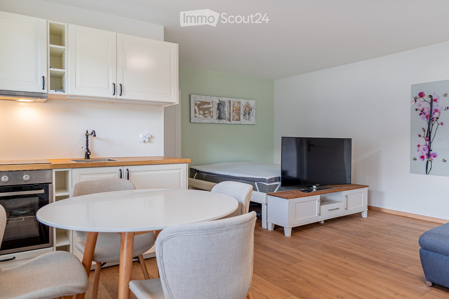 Open-concept studio apartment with white kitchen cabinets, wooden countertops, round dining table, gray upholstered chairs, TV stand, and hardwood flooring.