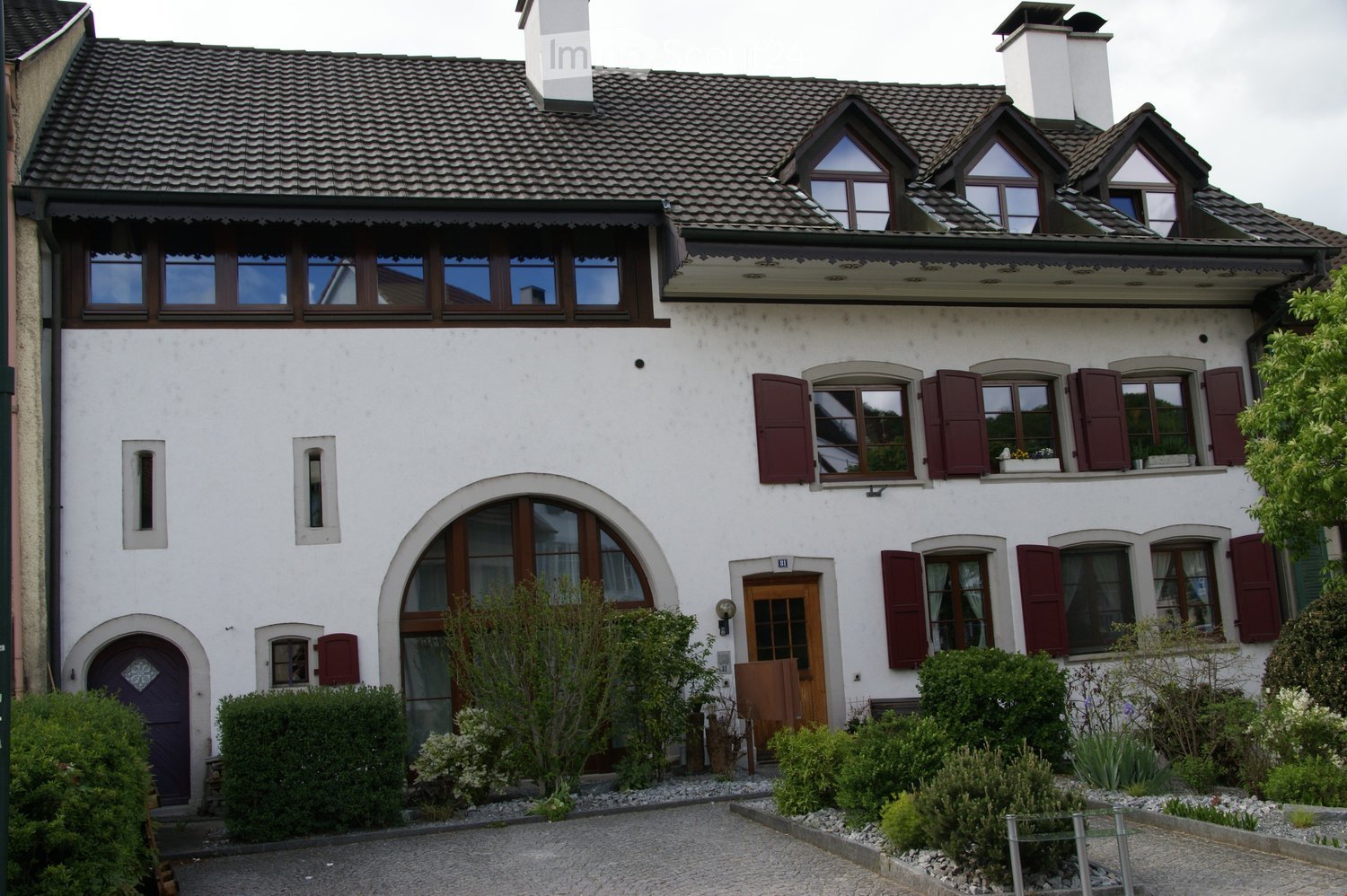 2-story house with a white exterior, arched windows, and red shutters. The roof has multiple dormers and chimneys. There is a small garden and landscaping in the front of the house.