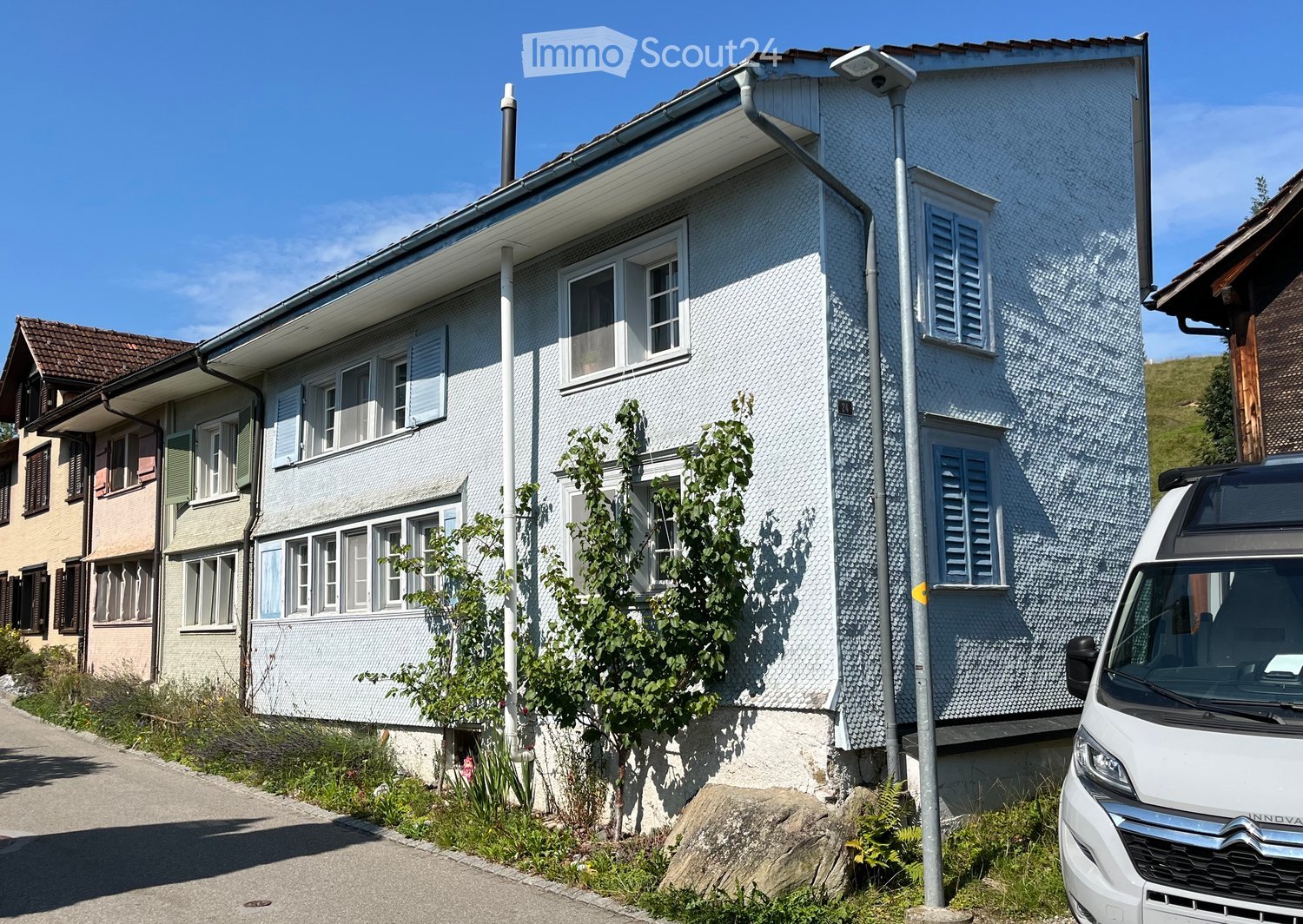 2-story residential building with white exterior walls, blue shutters, and a slanted roof. There is a parked car in the foreground.