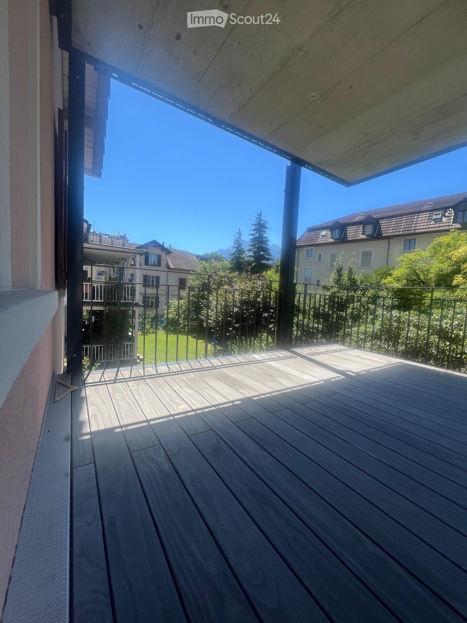 Spacious balcony with wooden flooring, overlooking a green garden area and surrounding buildings