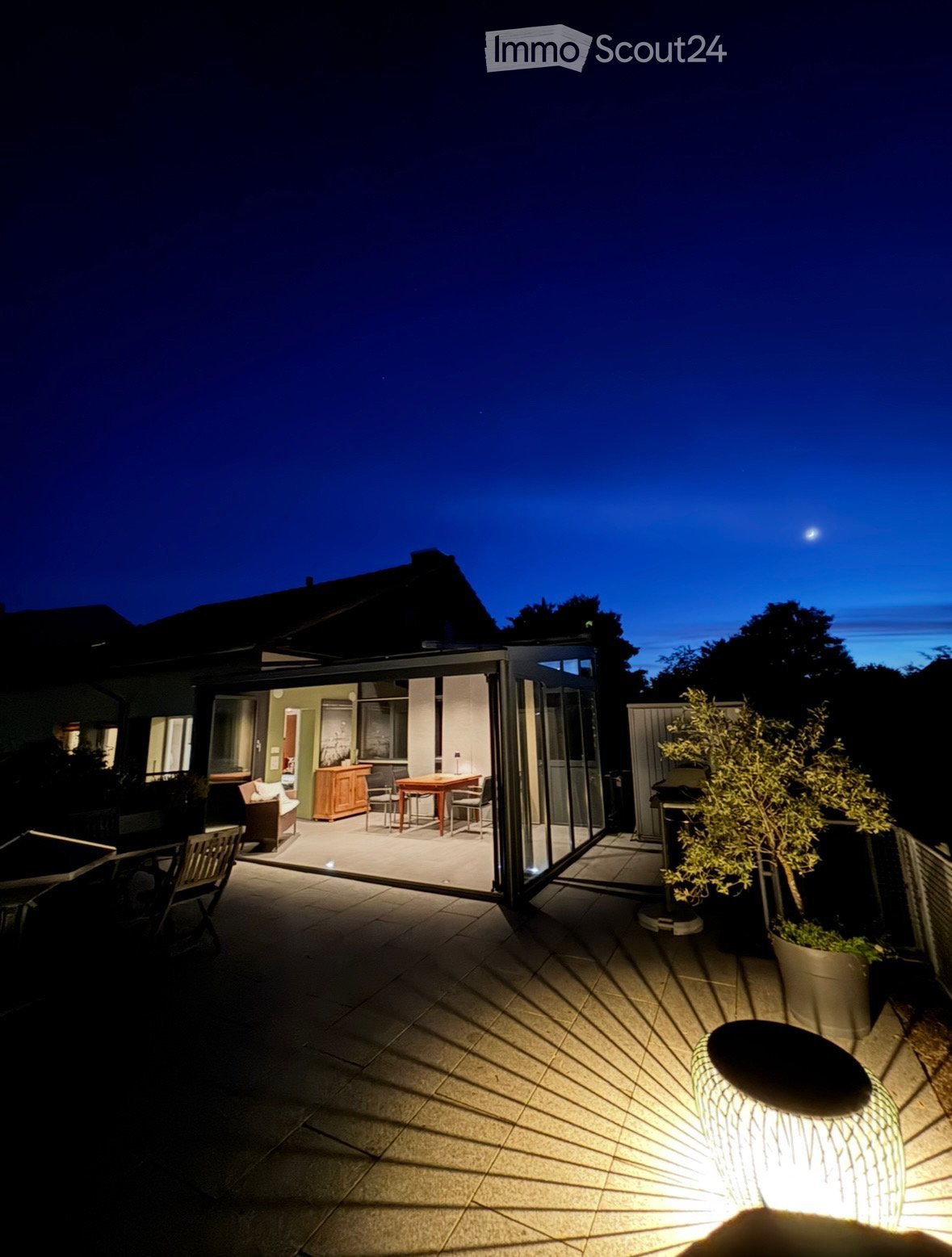 An outdoor living space with sliding glass doors, table and chairs, outdoor seating, tree, and decorative lighting. The moon is visible in the night sky.