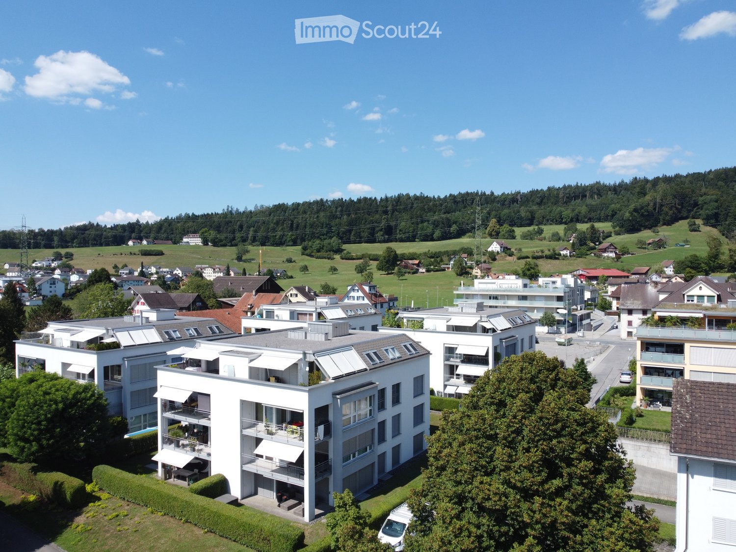 Aerial view of apartment buildings, surrounded by trees, grass, and a mountain. Solar panels on rooftops, balconies visible, a car parked, and a building with a brown roof.