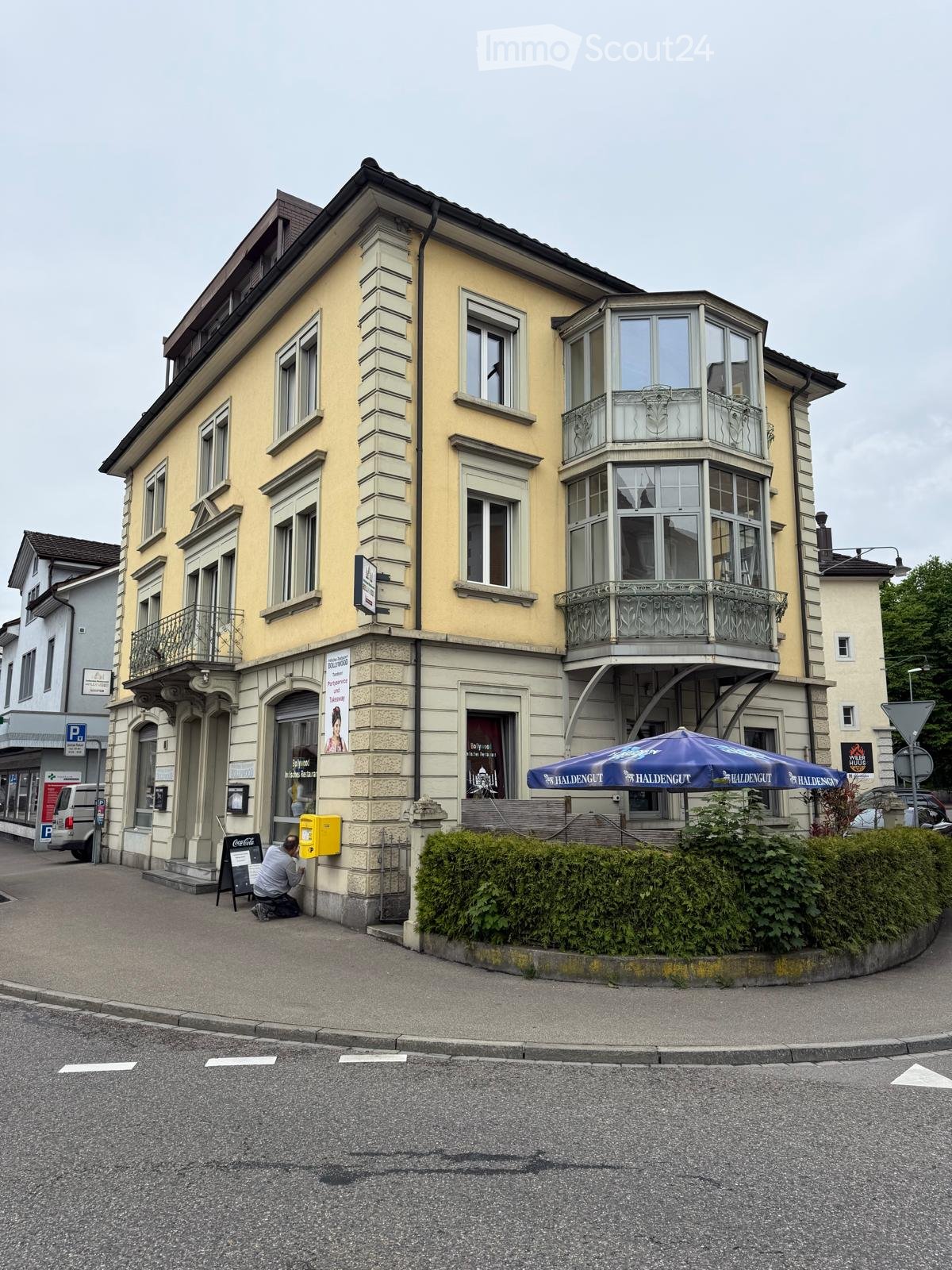 3 story building with yellow exterior, multiple windows, front has a sign and awning, bus stop is on the side of the street, green hedges on the right