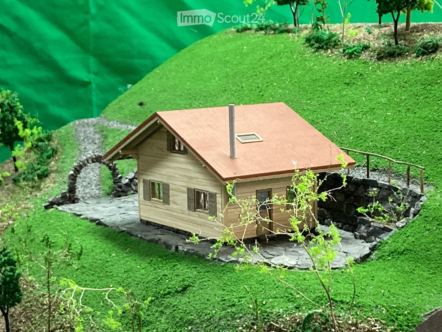 Miniature wooden chalet, red roof, surrounded by greenery, small front pathway, small waterfall, stone garden elements