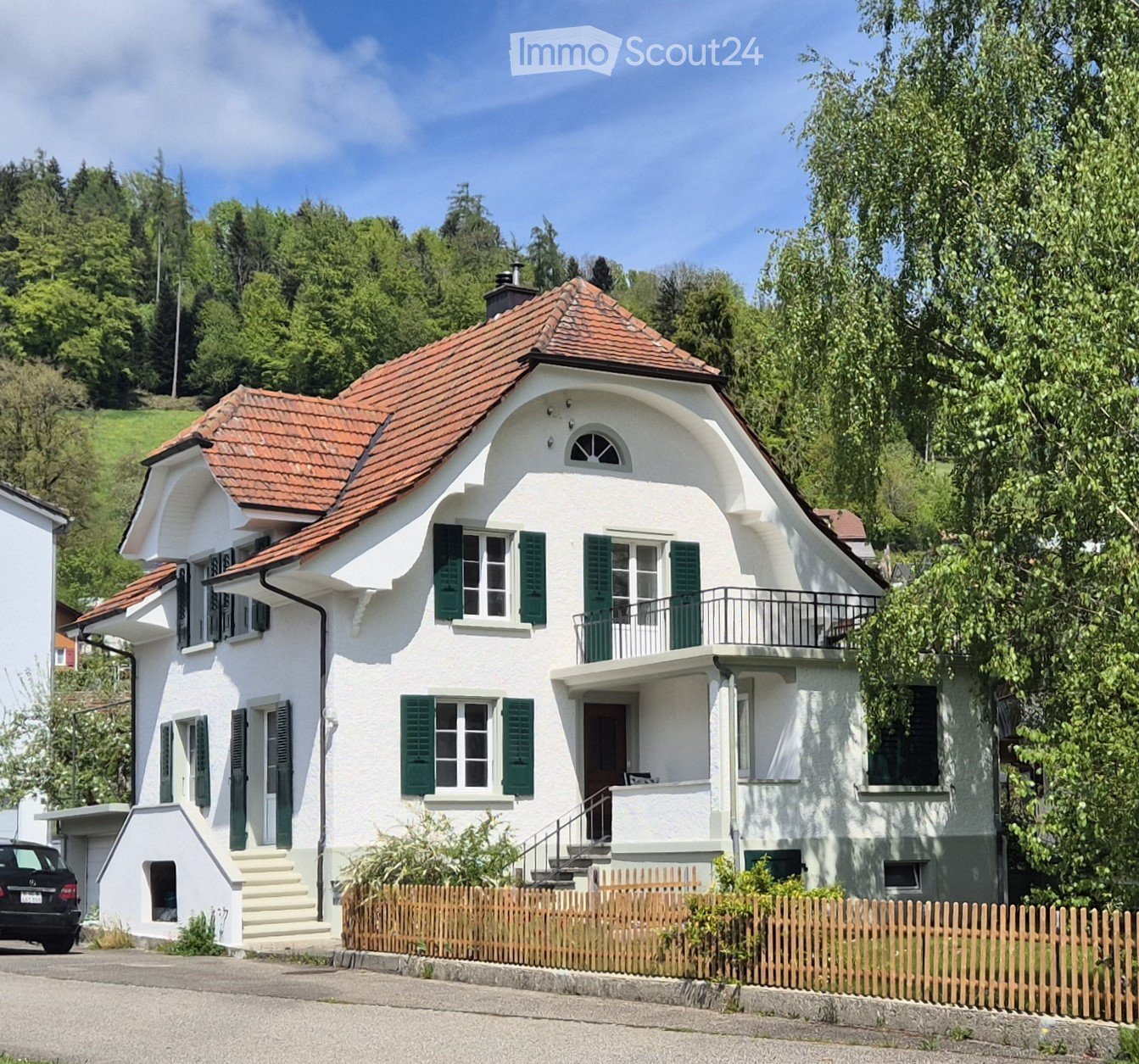 2 story house, white walls, red roof, green shutters, balcony, wooden fence, garden, black car parked, hillside in the background