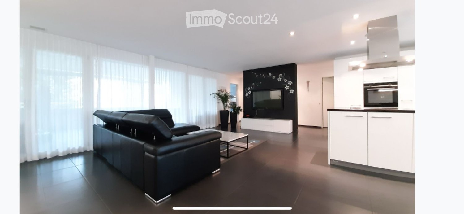 modern style, black sofa, white walls, gray flooring, glass coffee table, television mounted on wall, kitchen with white cabinets and appliances, potted plants, glass windows with curtains