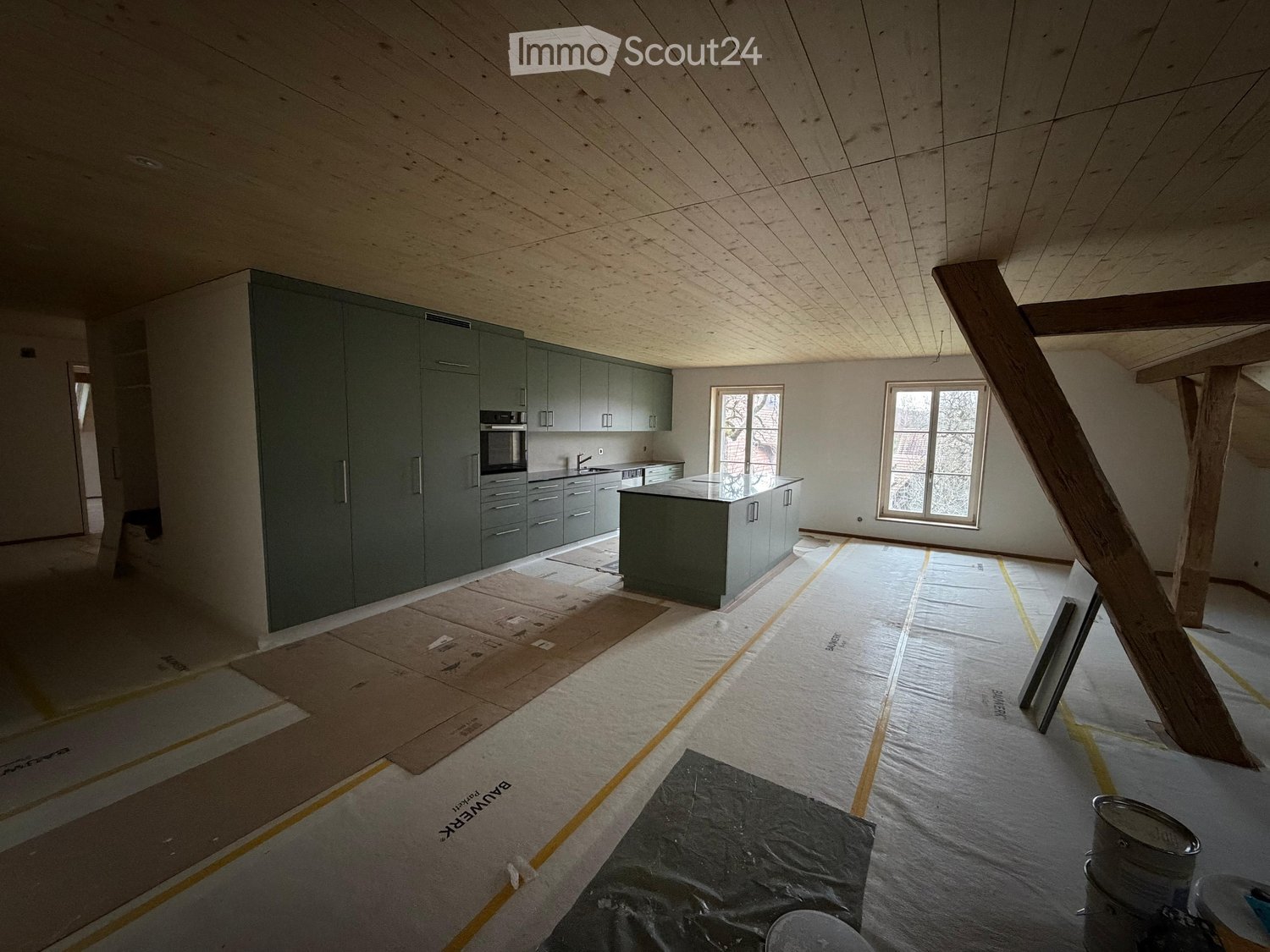 Under construction kitchen with modern cabinetry, central island, open ceiling beams, stainless steel appliances, unfinished floor, large windows, two wooden supports.