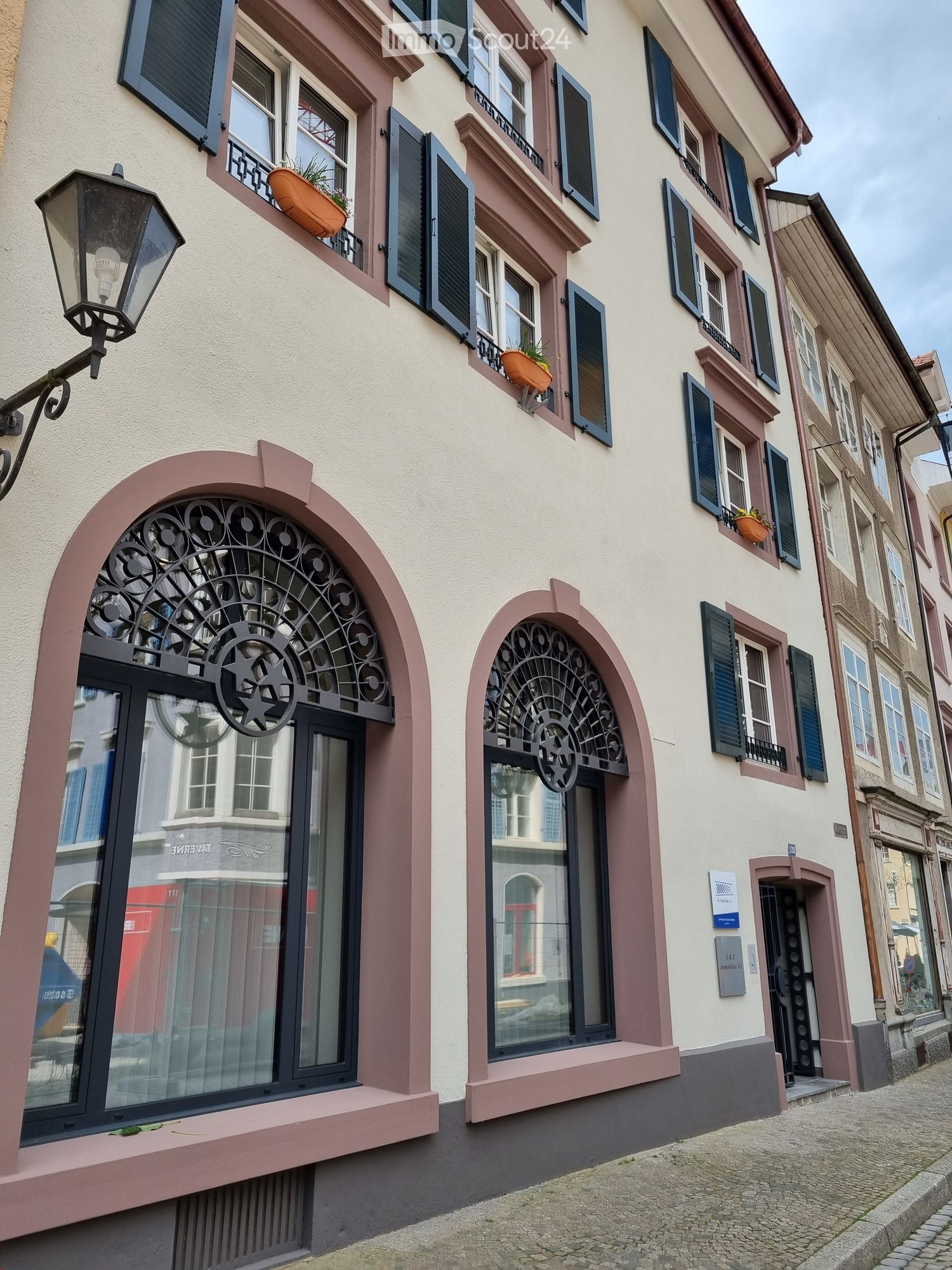 Urban facade, beige walls, three arched windows with black metal grilles, three balconies with potted plants, two lamps attached to the wall