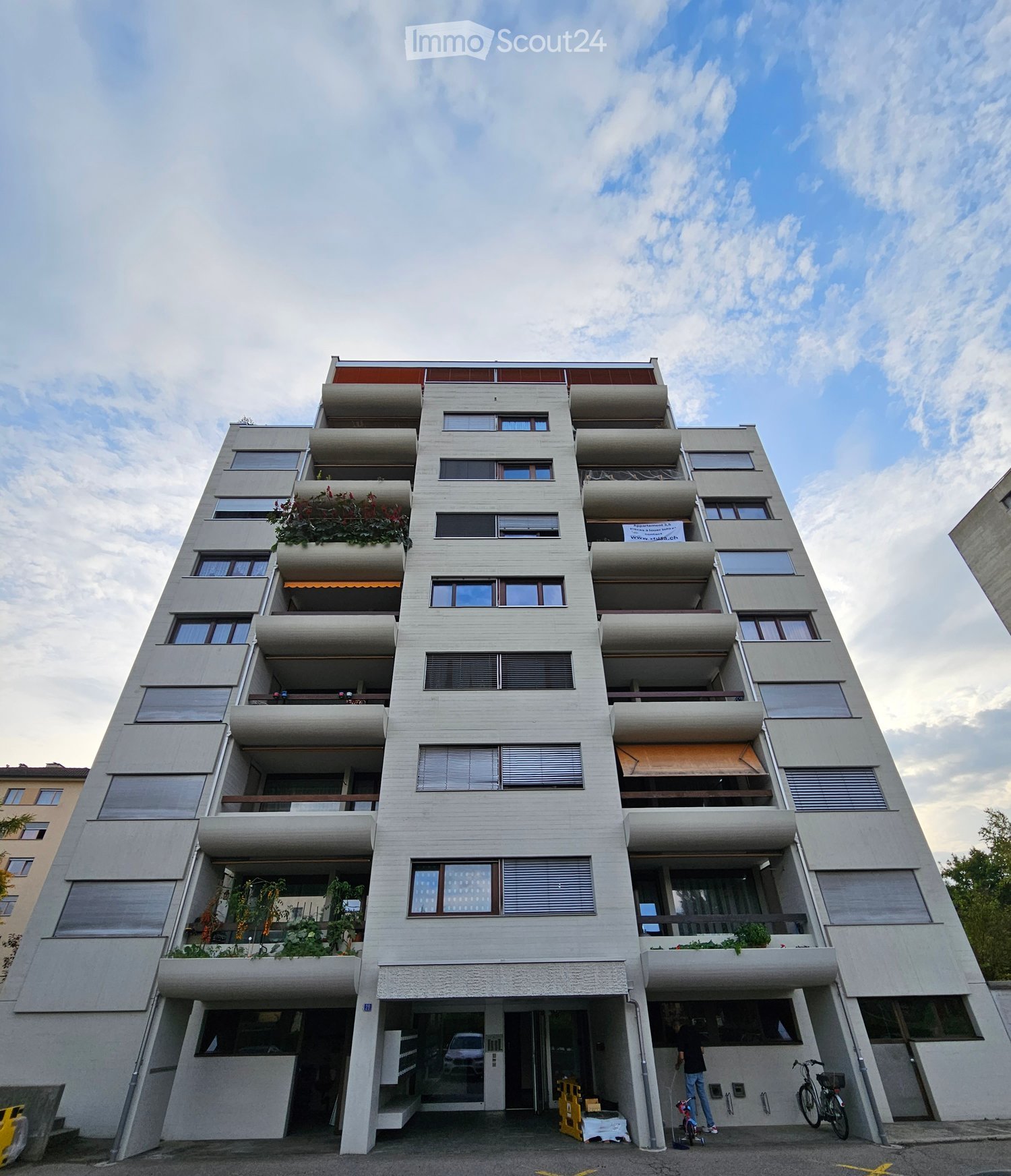Concrete multi-story building, balconies with plants, windows with blinds, entrance with garage and bike racks, sign indicating an apartment for sale on the top floor
