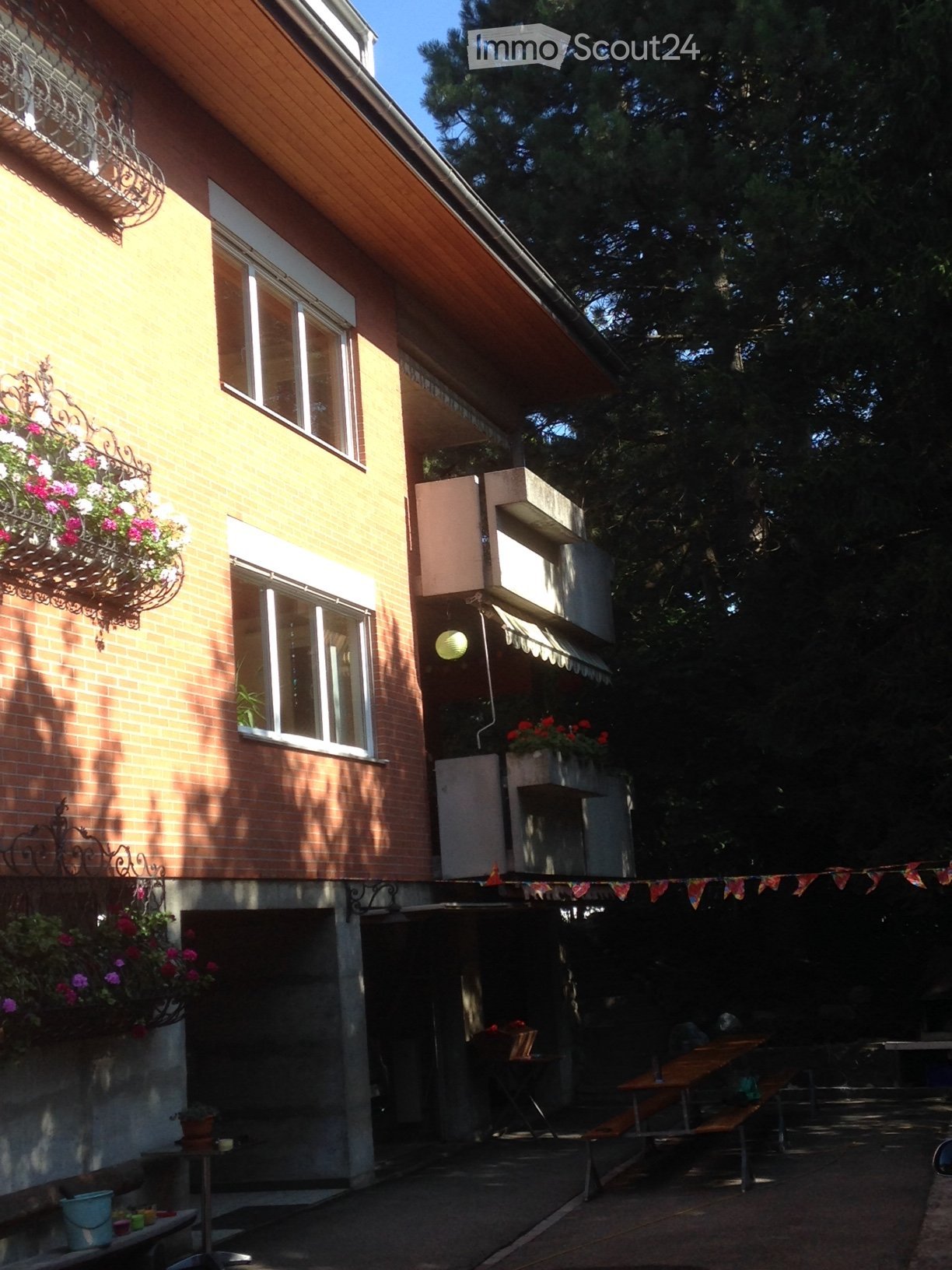 Exterior of the house with brick wall, balconies with flower boxes, a lamp hanging, and a garden with wooden benches and flags.