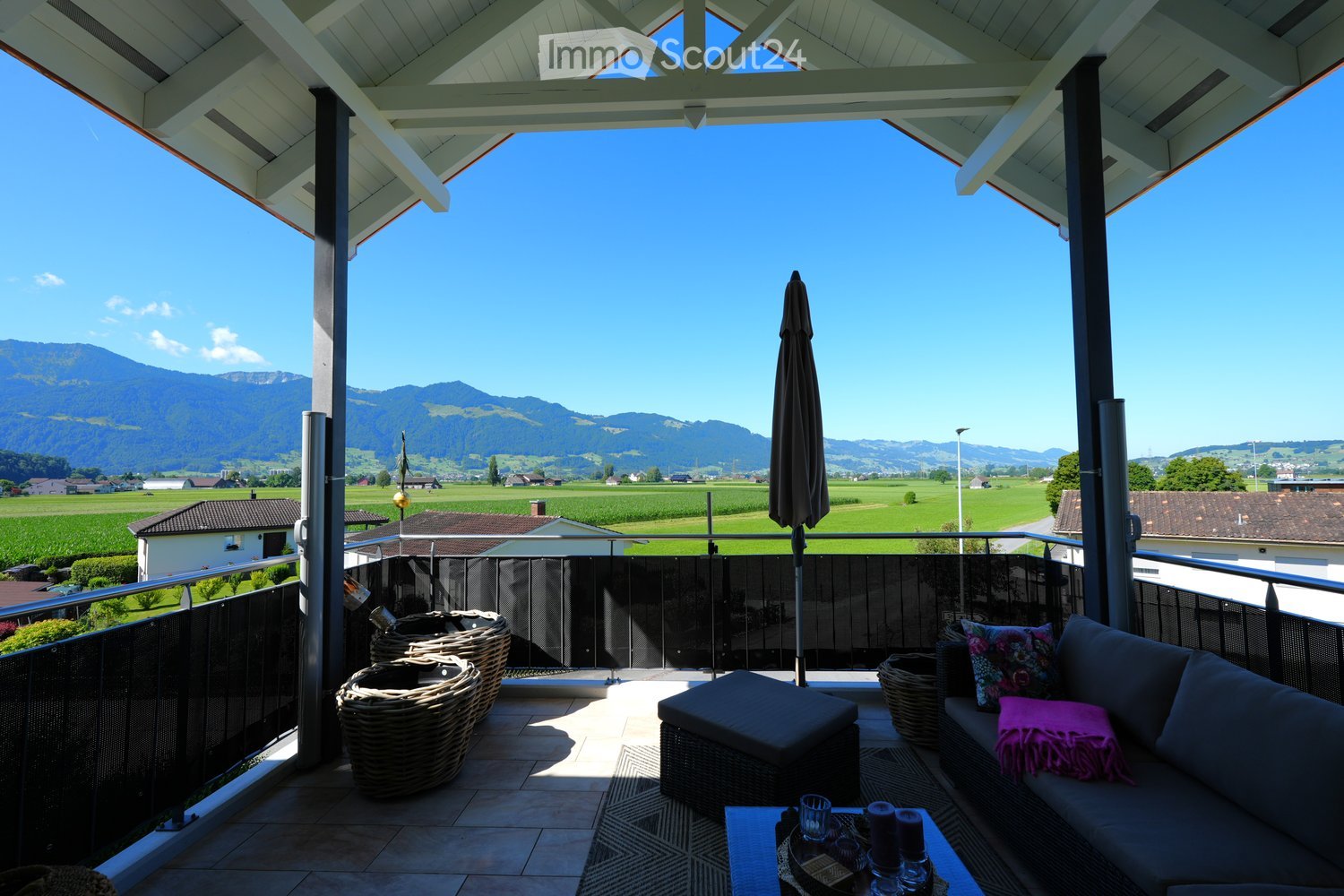 Balcony with railings, outdoor sofa with cushions and pillows, woven baskets, closed umbrella, tiled floor, table with glasses and candles, countryside view with mountains and fields