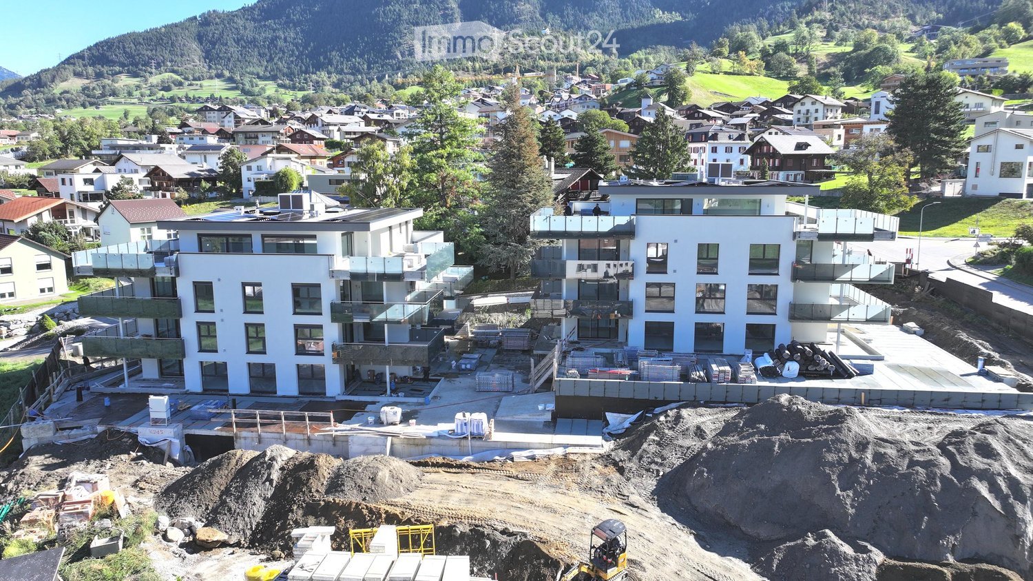 Aerial view of two modern apartment buildings under construction with terraces, surrounded by trees and a mountainous backdrop.