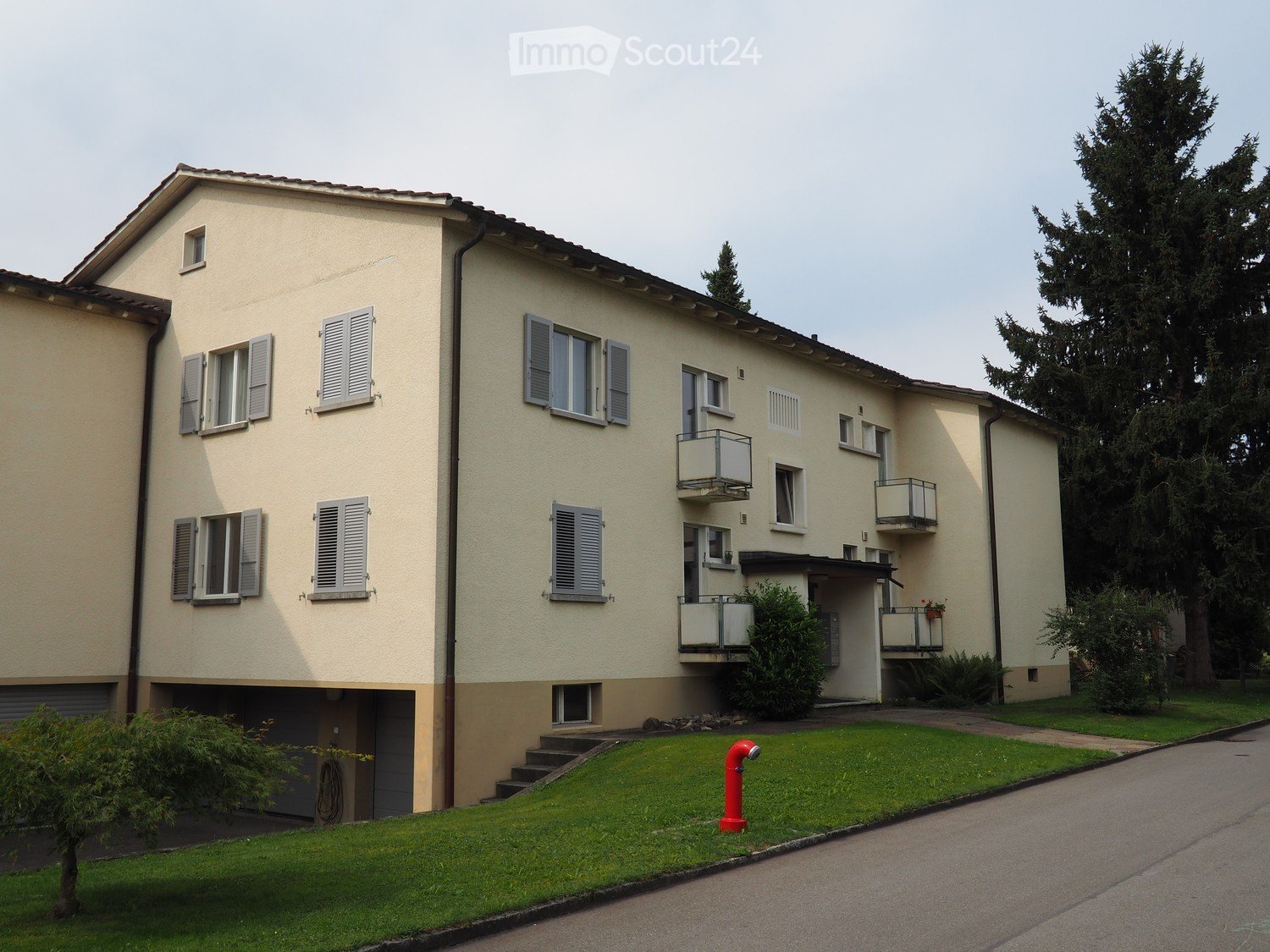 Three-story building with beige walls and white shutters. Each floor has several windows and balconies. Grassy area with plants in front and stairs leading up to the building.