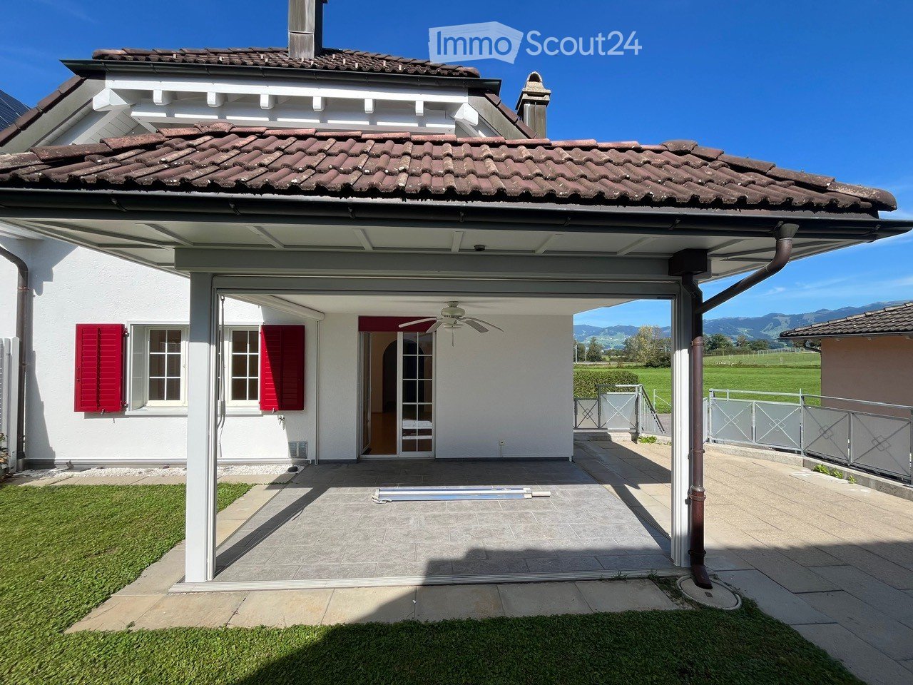 White house, brown roof, red shutters, terrace with ceiling fan, garage, lawn, fenced area, green field, mountains