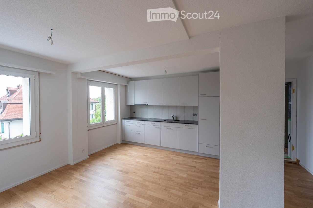 empty room, white walls, wooden floor, large windows, white kitchen cabinets, modern kitchen with black countertop, sink, faucet, open doorway