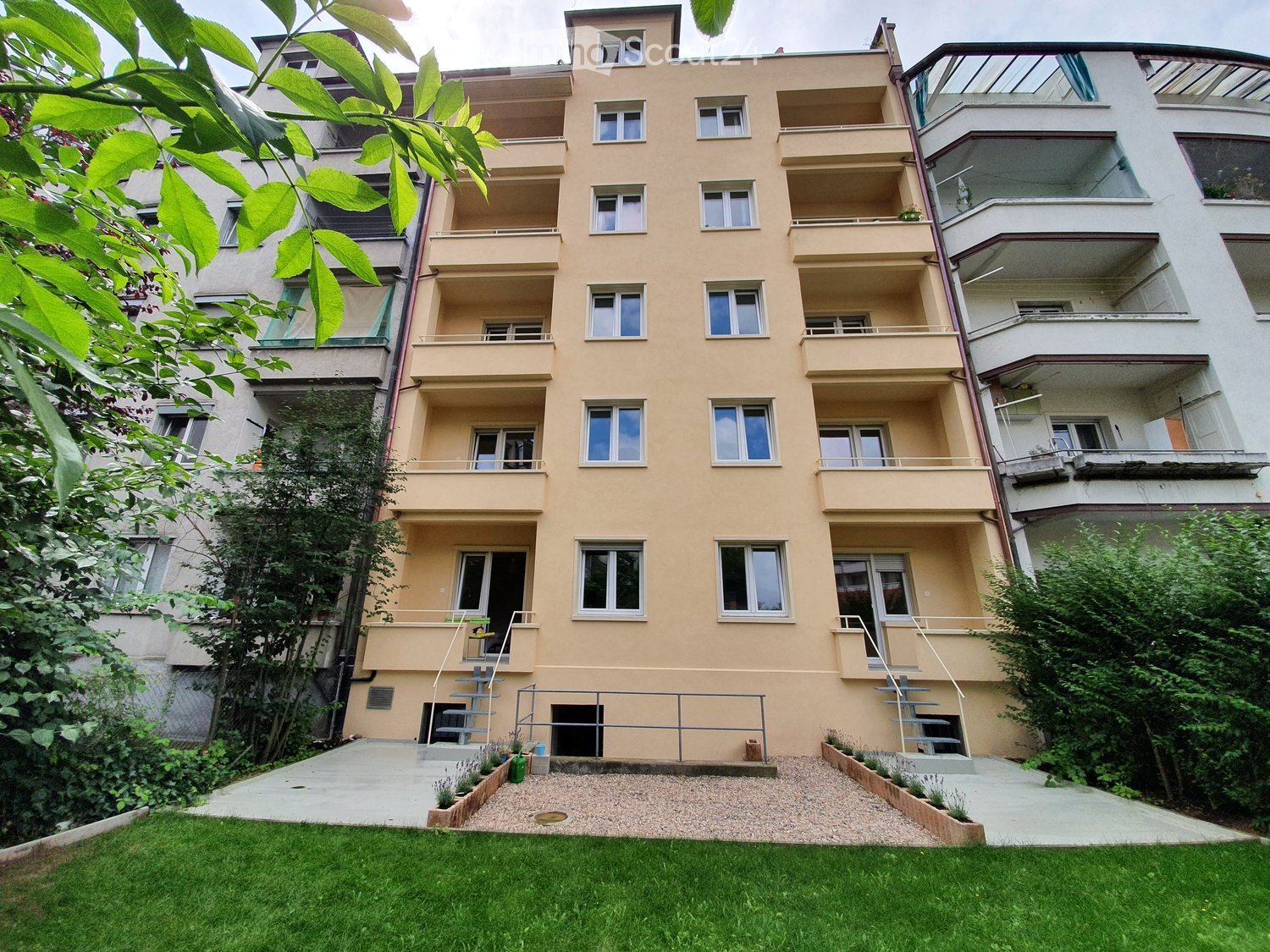 4 story building, yellow walls, windows, balconies, front yard with gravel and plants, 2 entry stairs, fence, other buildings in background