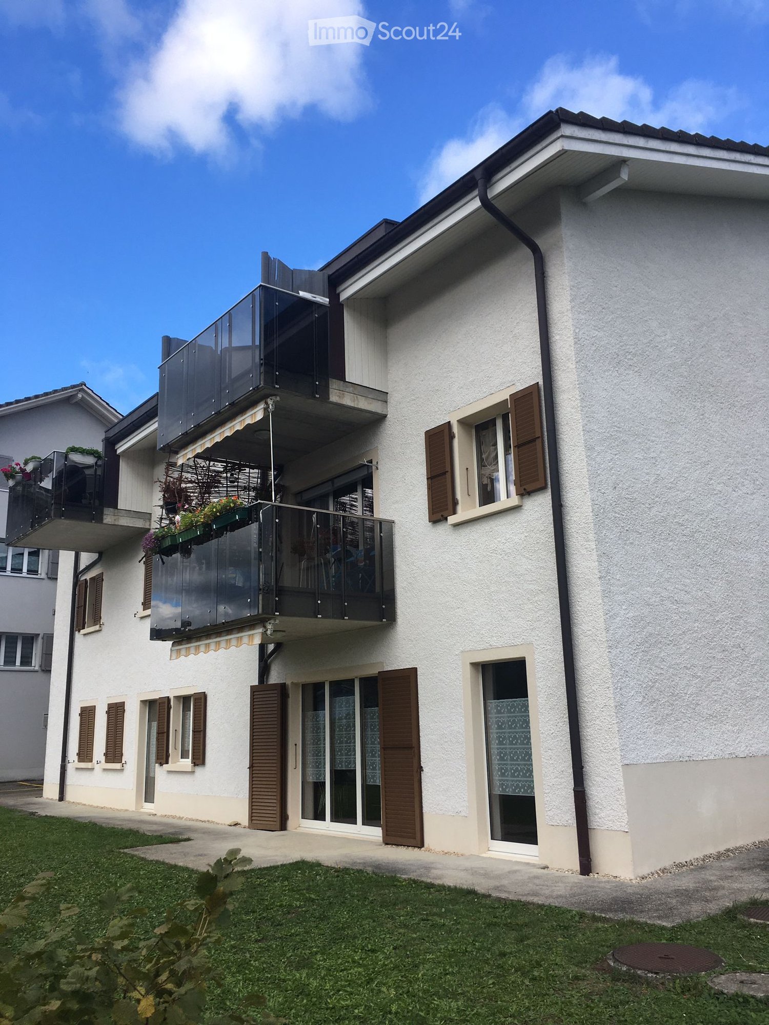 Two story apartment building with balconies, white exterior, brown shutters, glass sliding doors, grassy area in front