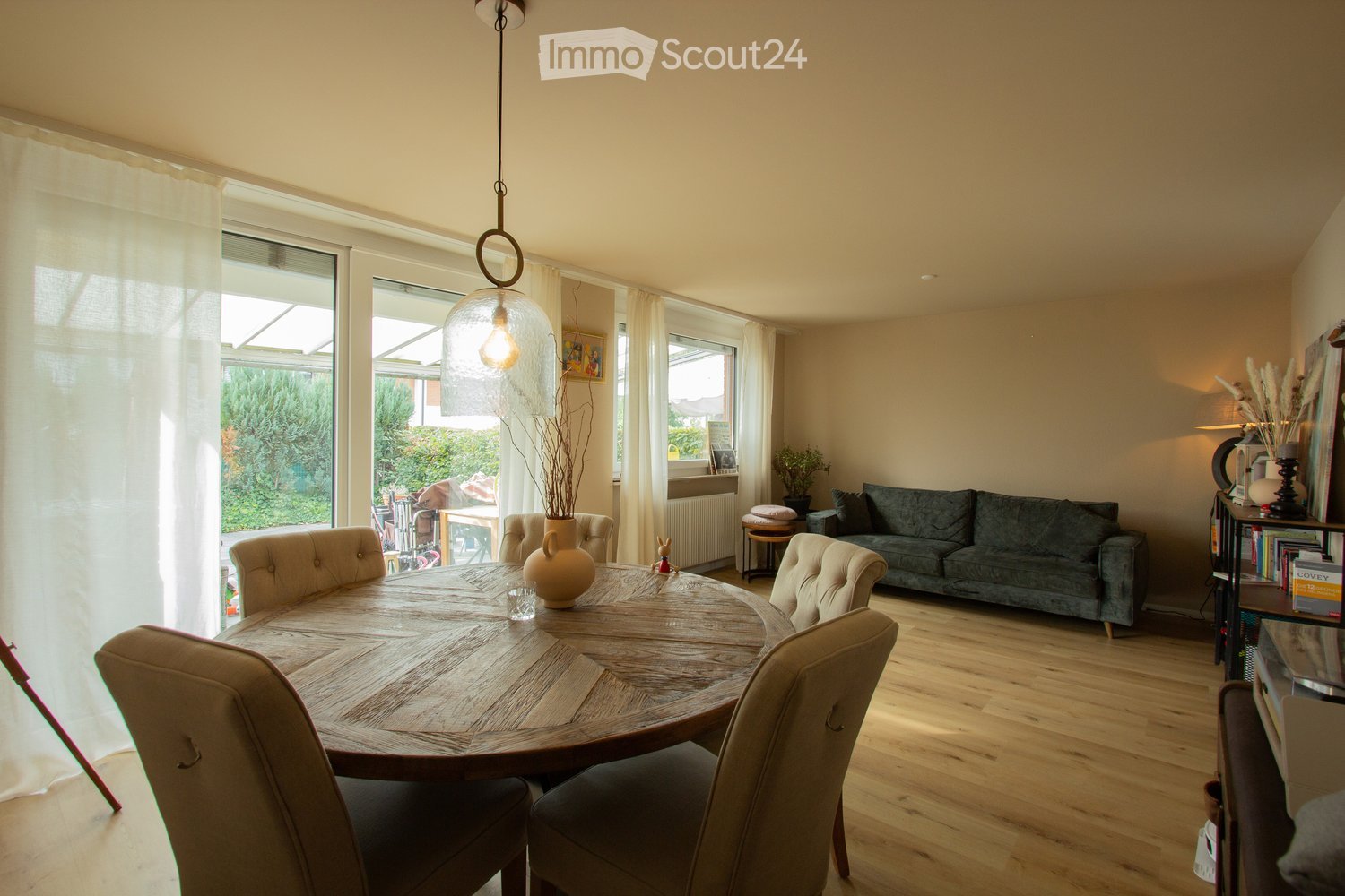 Round dining table, four chairs, wooden floor, glass doors leading to balcony, couch in corner, large windows