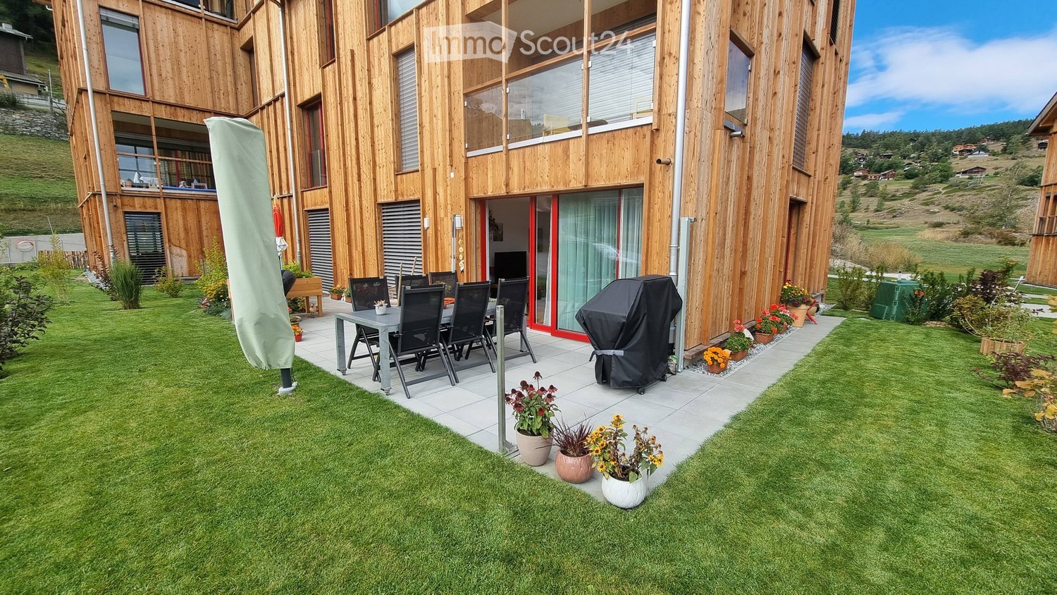 Modern wooden building, dining table and chairs on terrace, multiple potted plants, green lawn