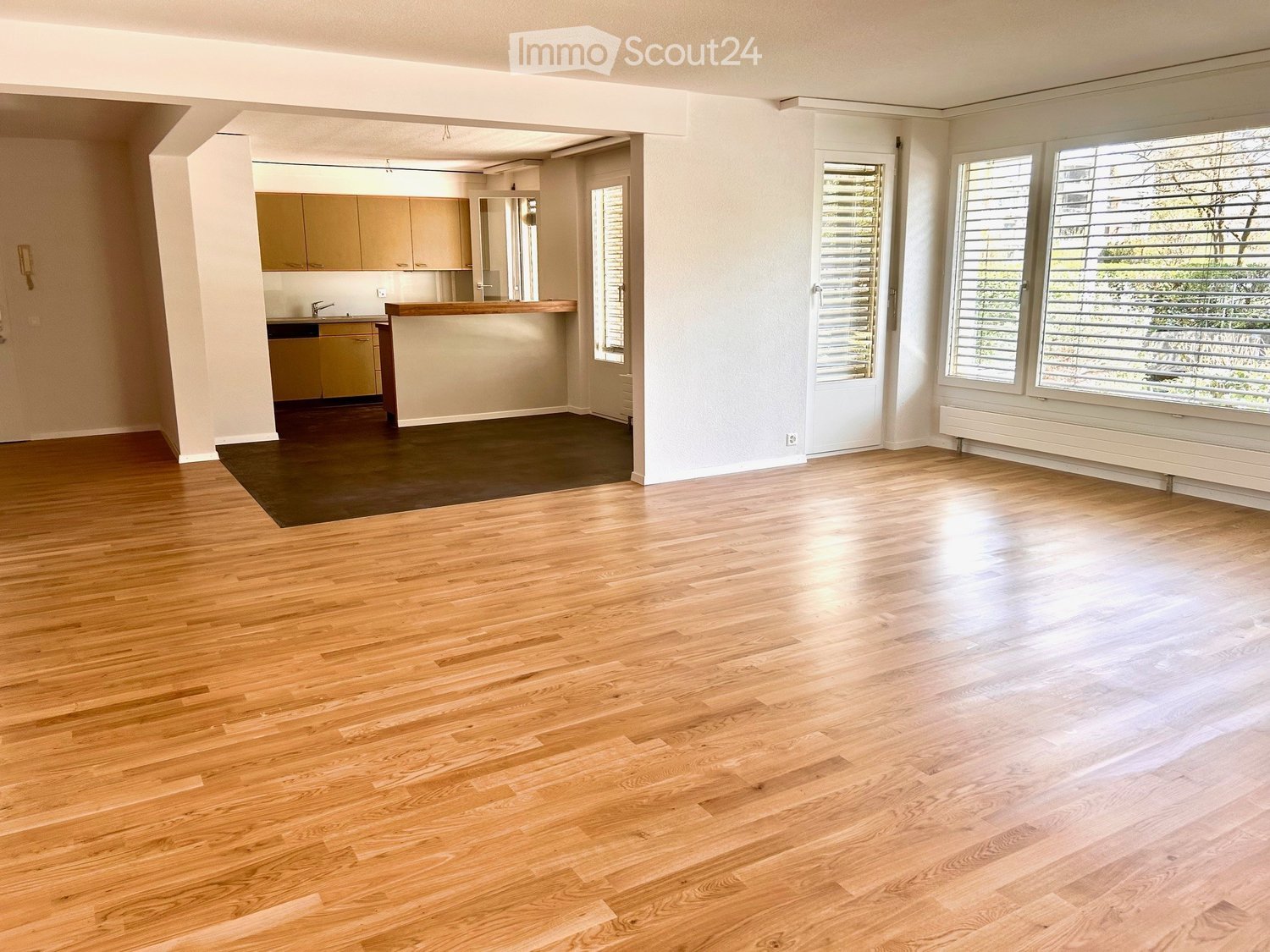 The image shows a spacious apartment with hardwood floors, large windows, and an open floor plan. The living area has a wooden floor and leads into a kitchen area with wooden cabinets. The apartment appears to be well-lit and has a modern, minimalist desi