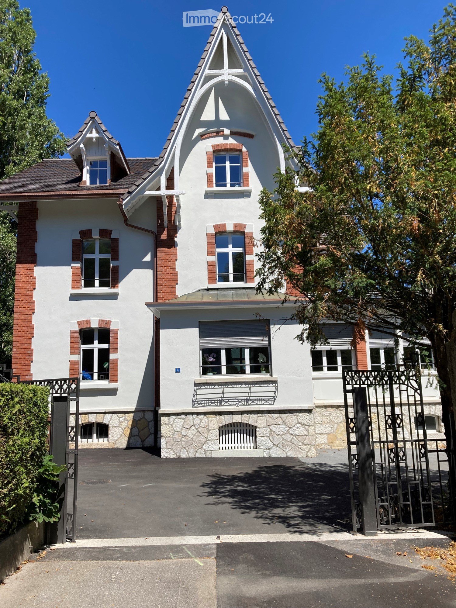Two-story house, white painted exterior with red brick accents, arched windows, large front yard, decorative wrought iron gate, large tree in front, paved driveway
