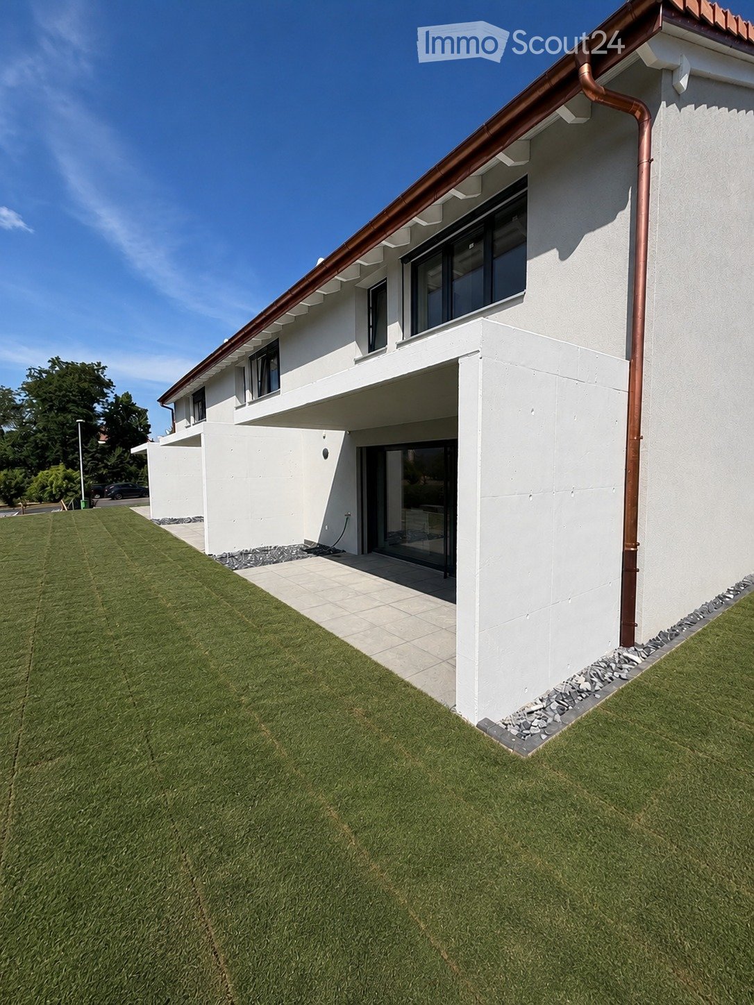 Modern two-story house with white exterior, large windows, and a covered entrance. The house features a well-maintained lawn and a paved pathway leading to the entrance.