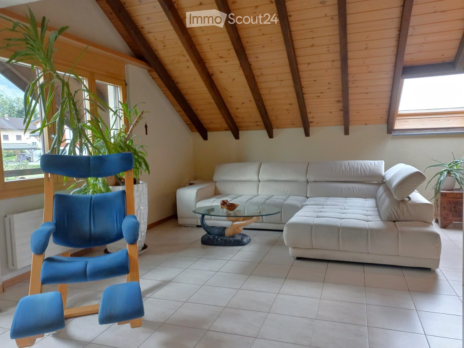 a living room with a sectional sofa, glass coffee table, wooden ceiling, blue high back chair, and potted plants
