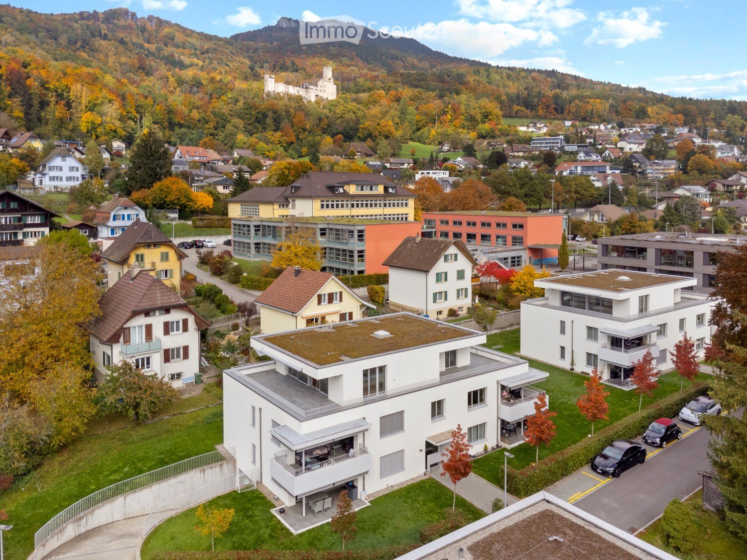 Modern 4-storey terraced house, white facade, green roof, multiple balconies, surrounded by other houses and greenery, clear view of the mountains and village