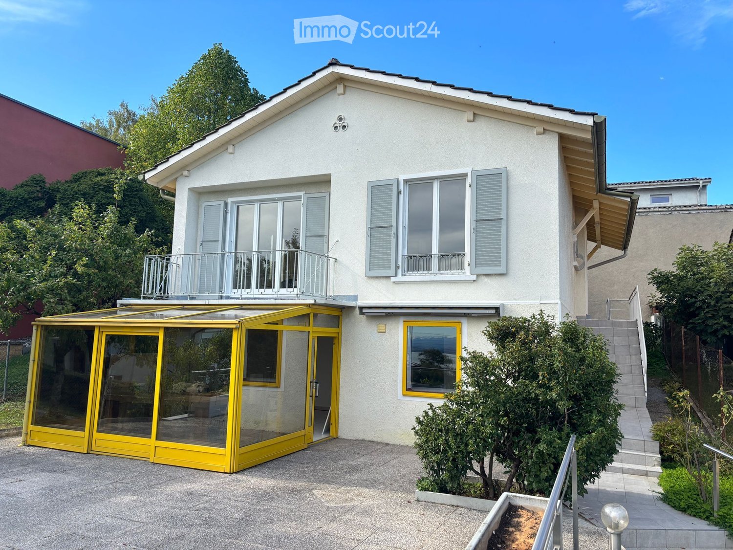 two-story house, white exterior, yellow-framed windows, yellow glasshouse, balcony on the second floor, driveway with a glass carport