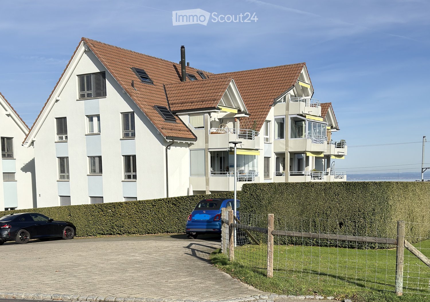 multi-story residential building, brown roof, solar panels, white exterior, balconies, two cars parked