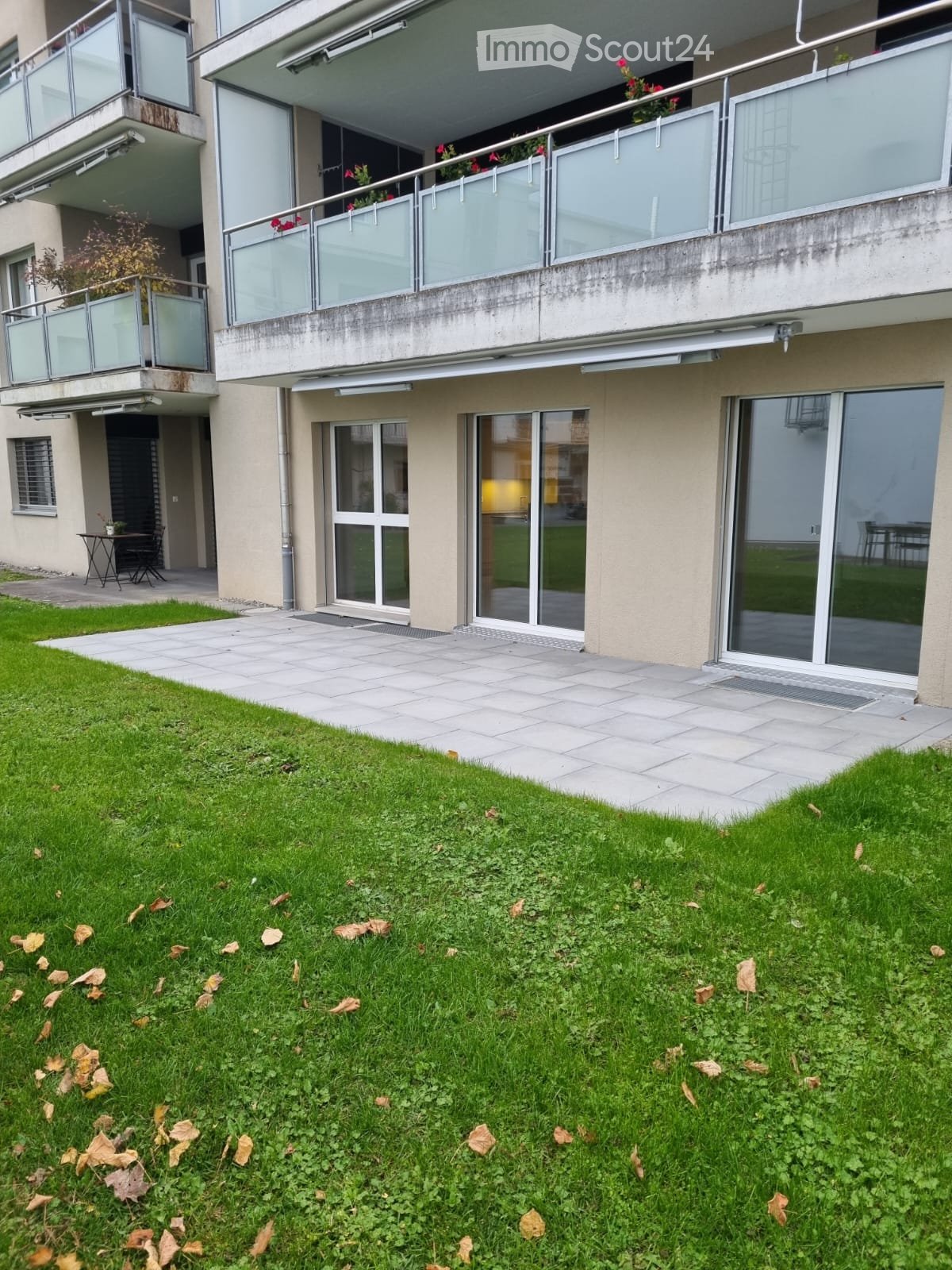 Two-story building, large glass windows, glass doors, paved walkway, balcony with plants, well-maintained grassy lawn