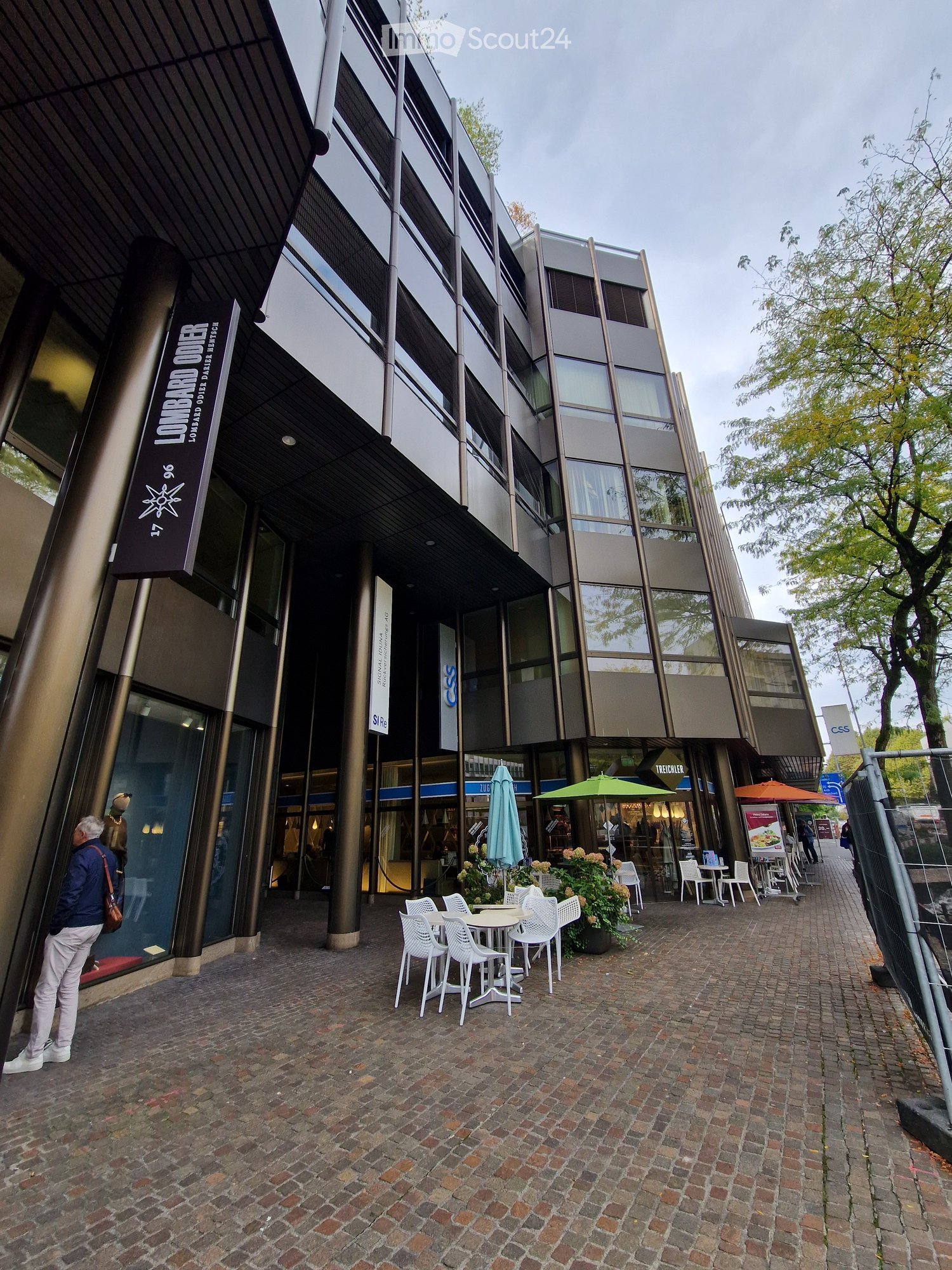 Modern building with glass facade, large entrance, cafe-style seating area outside, brick flooring.
