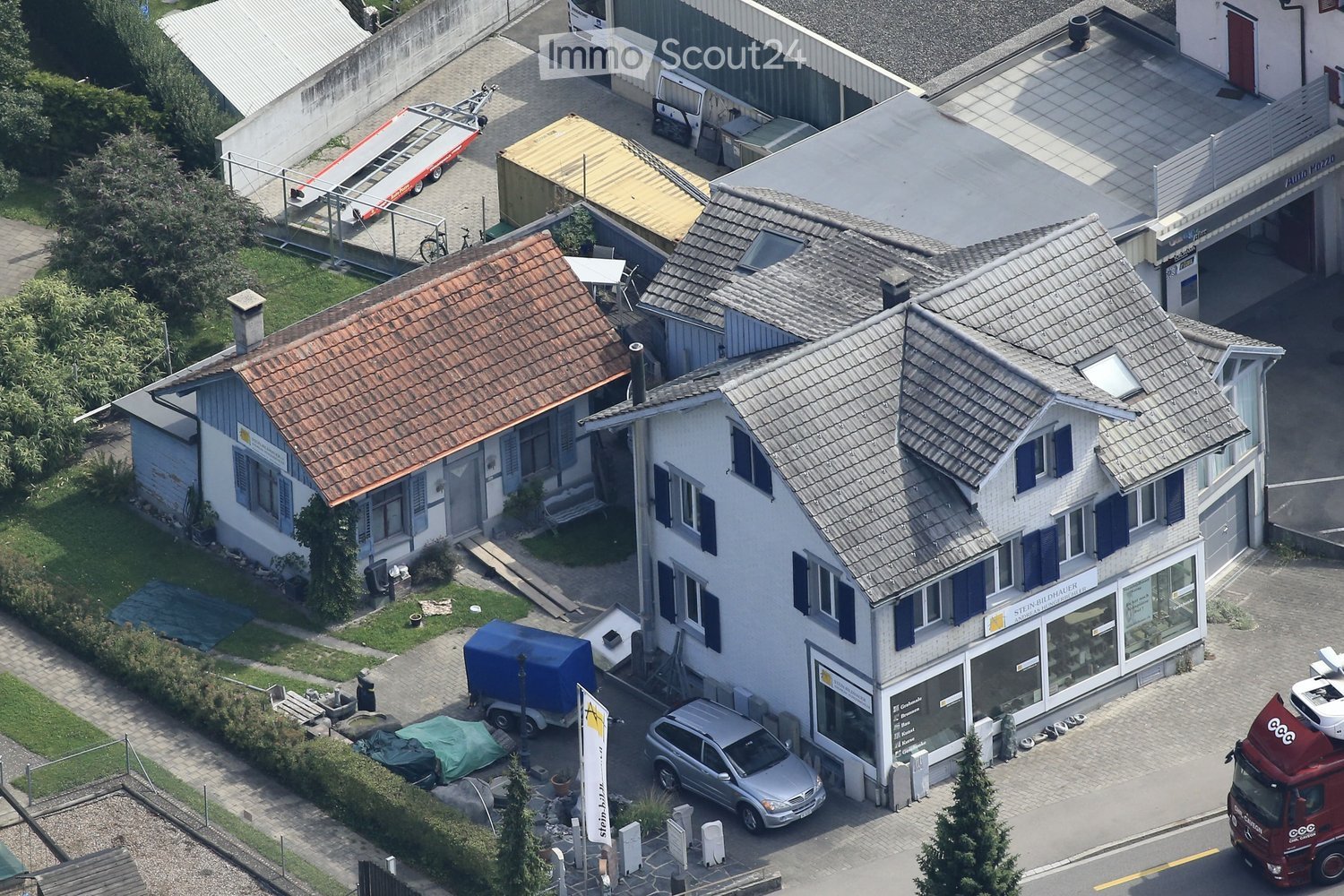 Two story house with red roof, blue shutters, attached building with windows, parked car, green bush, pathway, street, red truck