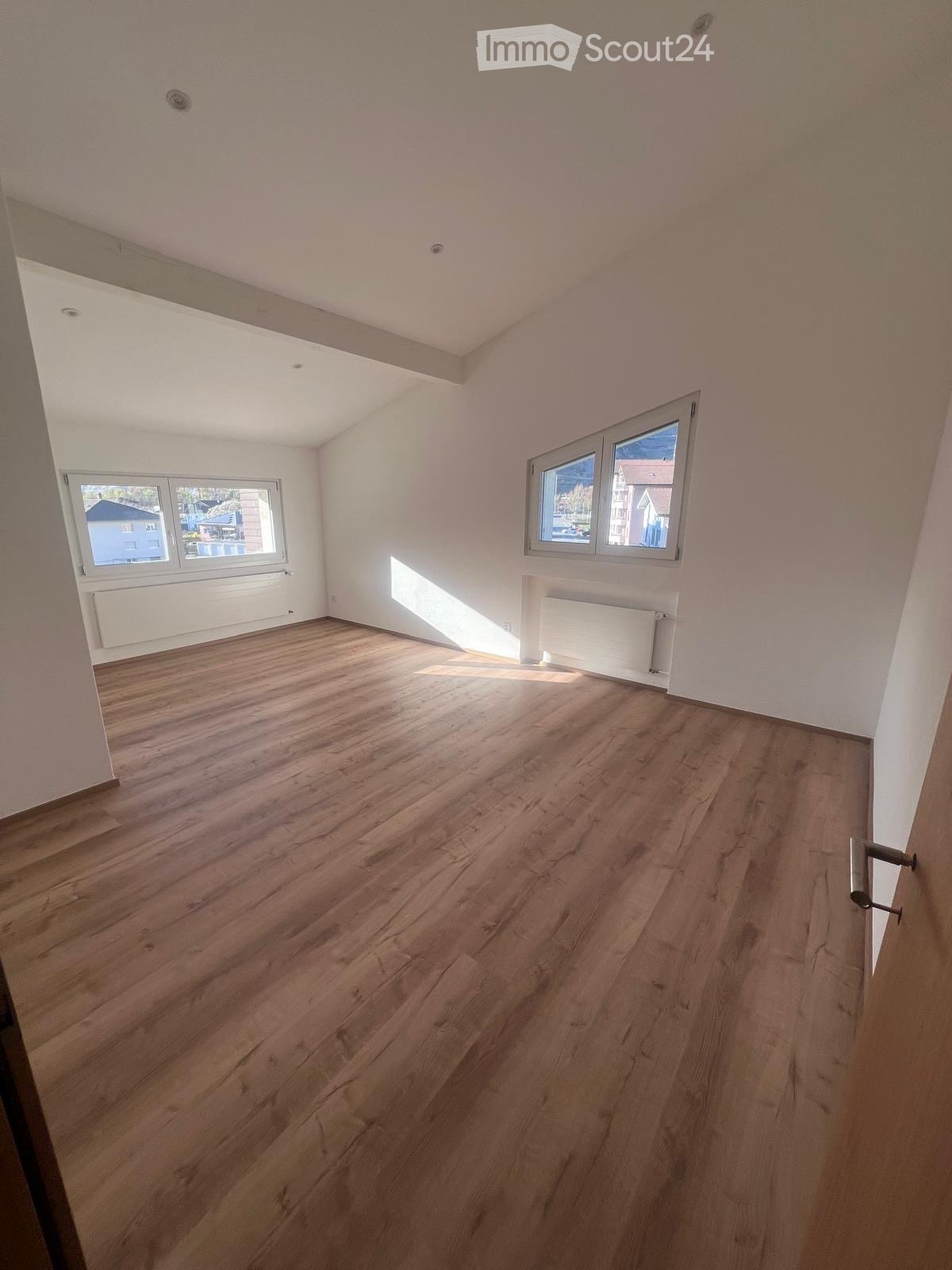 Spacious room with hardwood flooring, white walls, and large windows providing natural light. The room appears to be a living or bedroom area.