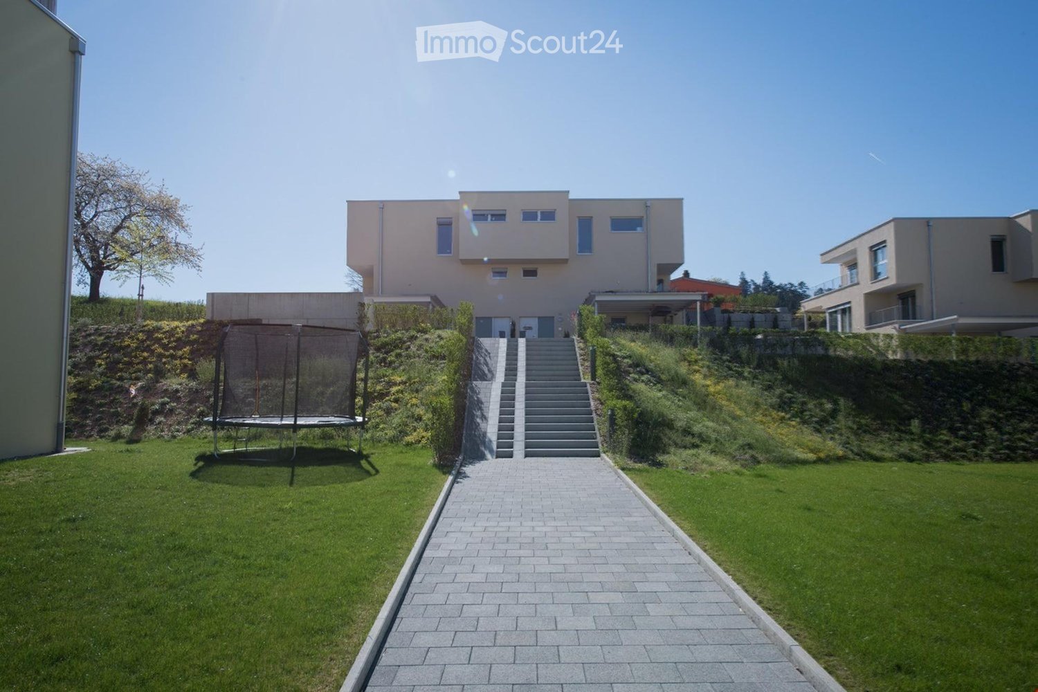 2-story modern house, beige facade, large windows, driveway leading to house entrance, lawn with stairs leading up to the house