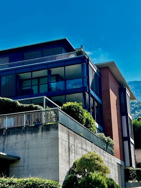 three-story building with blue trim, concrete foundation, green shrubbery, and glass balcony railings