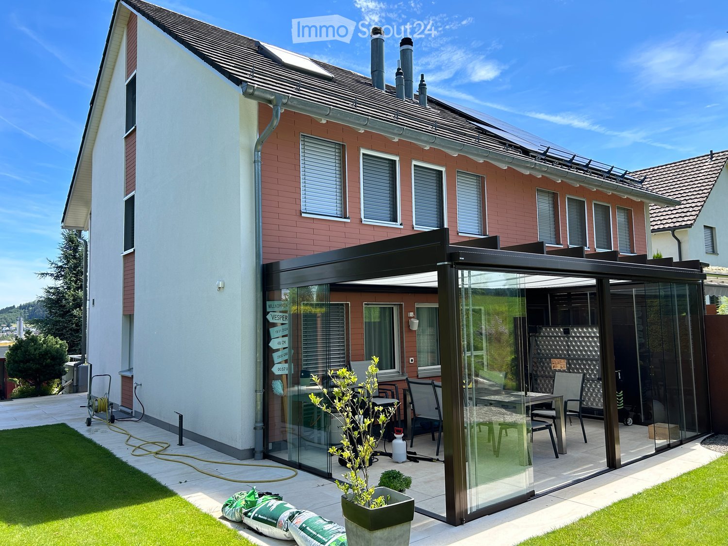 Two-story house, brick walls, solar panels, enclosed glass terrace, dining table, outdoor furniture, grassy lawn