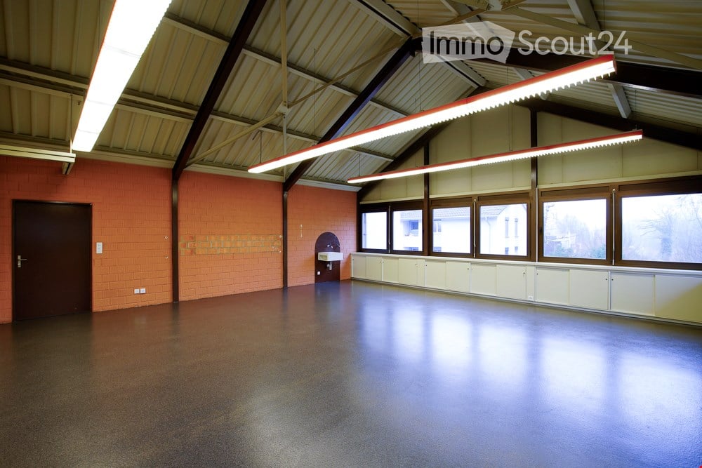 Large room with high ceiling, brick walls, fluorescent lighting, windows, and wooden-framed doors.