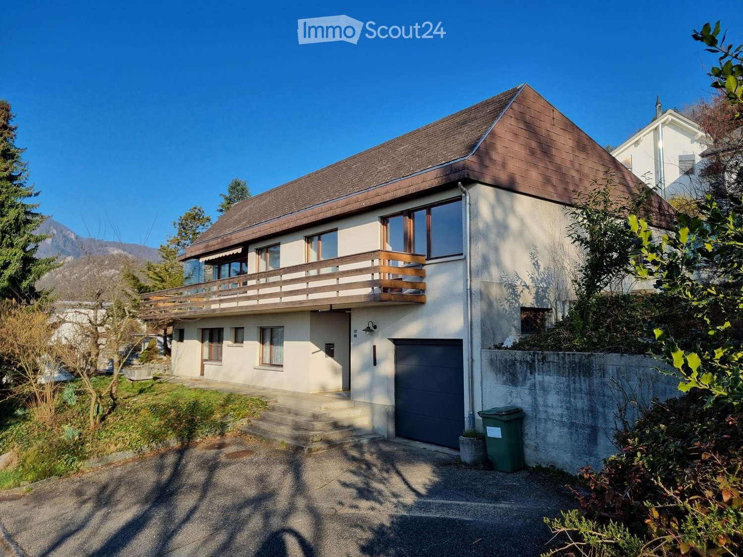 2-story house with a slanted brown roof, white exterior walls, and a balcony on the upper level. The house has a garage and is surrounded by trees and greenery.