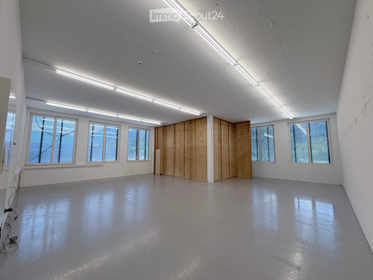 Empty room with white walls, large windows, and a wooden partition in the center. The floor is smooth and gray, and the ceiling has exposed lighting fixtures.