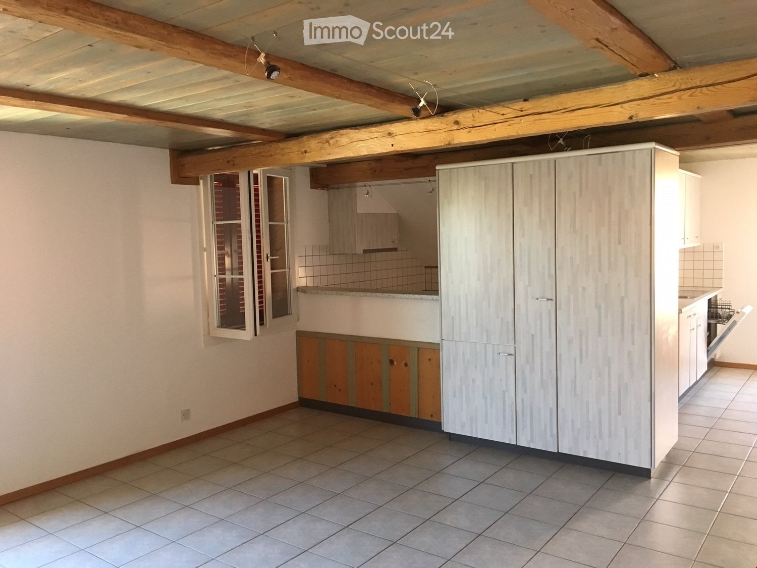 Empty kitchen with wood ceiling, white walls, wooden cabinets, large tiles, stainless steel sink, window with wooden frame