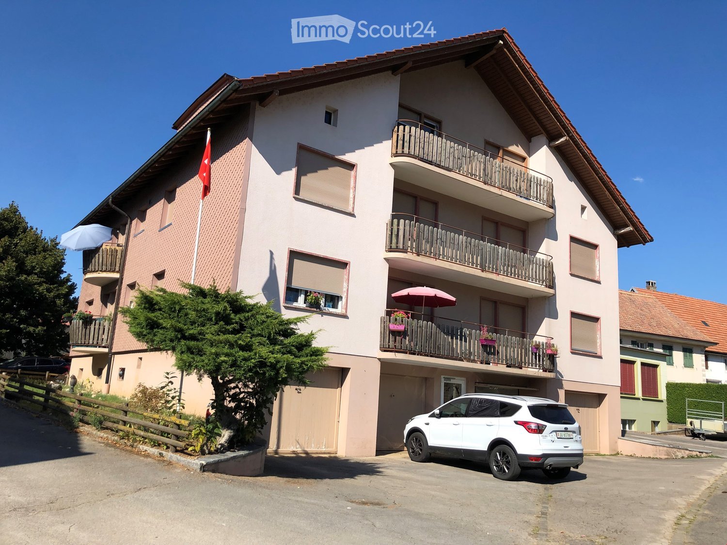 Three story apartment building, beige painted, red roof, several balconies with flowers, white SUV parked in front