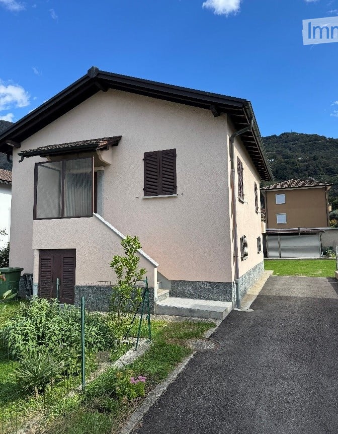 Two story house, concrete exterior, red roof, large windows, balcony, driveway, small garden, neighboring house in the background