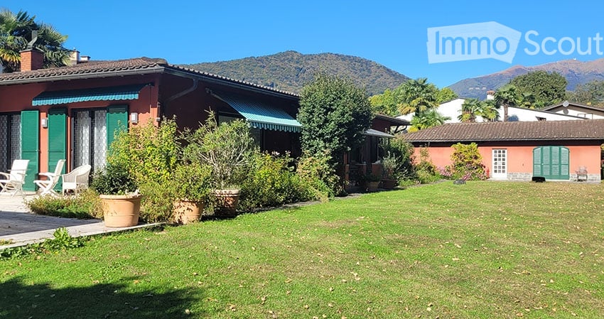 Red brick house with green windows and awnings, patio with two chairs, lush garden with various plants and trees, green lawn
