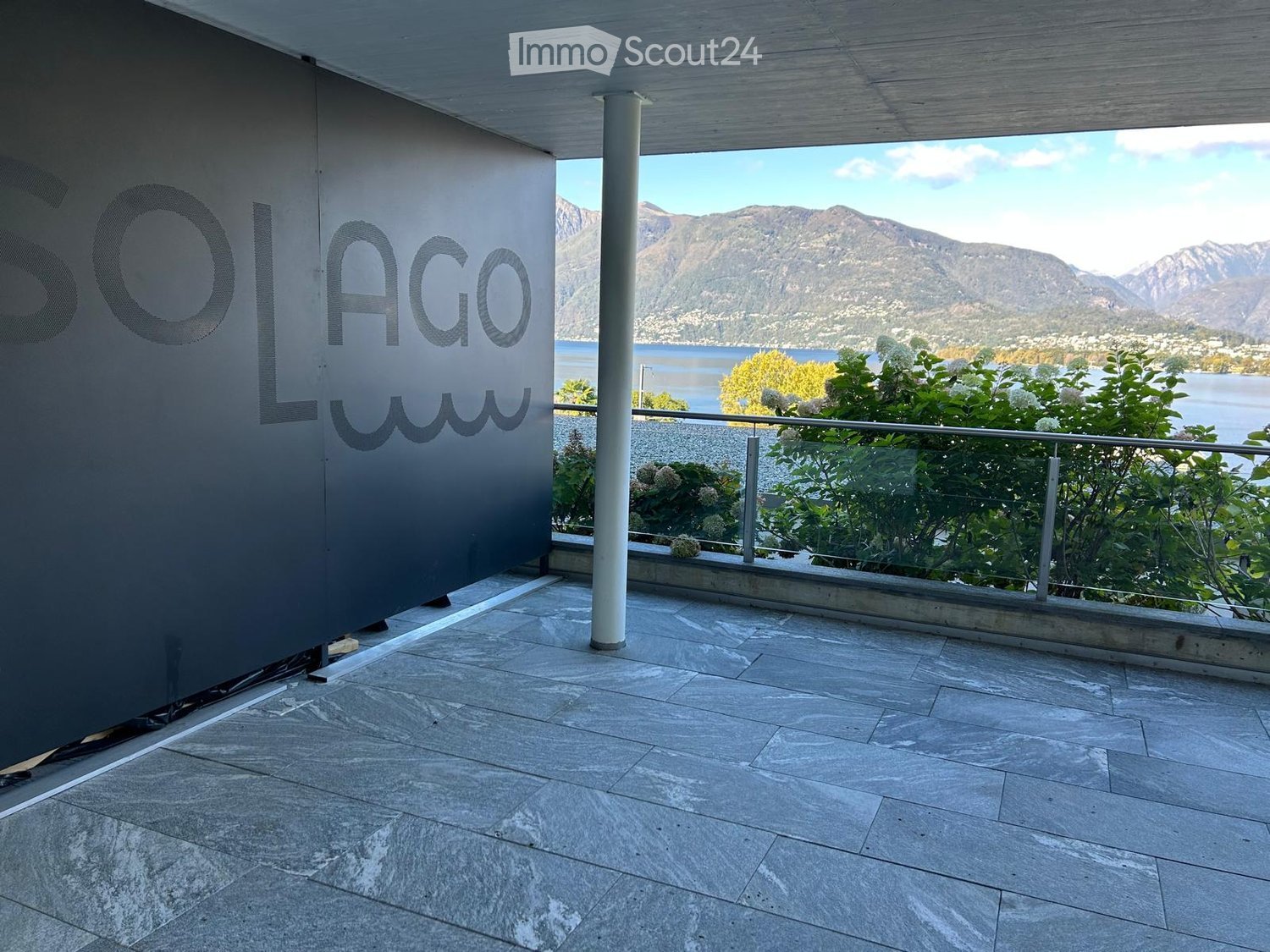 Outdoor terrace with grey tiled floor, black wall with 'SOLAGO' written, and a glass railing overlooking a lake and mountains.